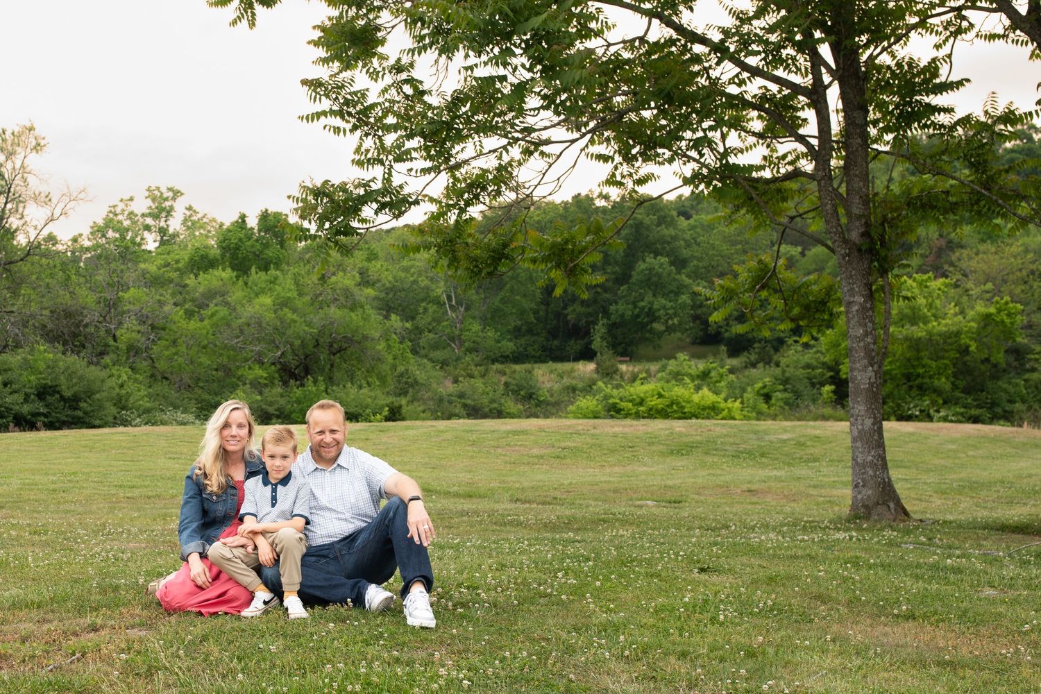 Family of three sitting together on green grass near a large tree in a scenic outdoor park setting.