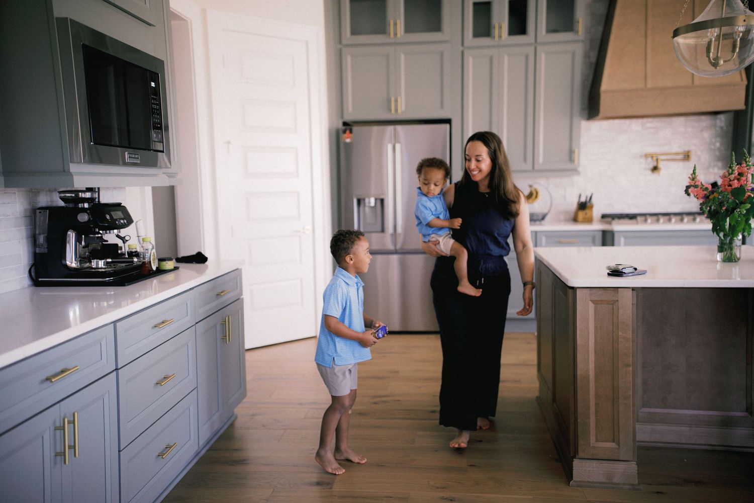 Woman in black dress holds toddler while older child plays in modern gray and wood kitchen.