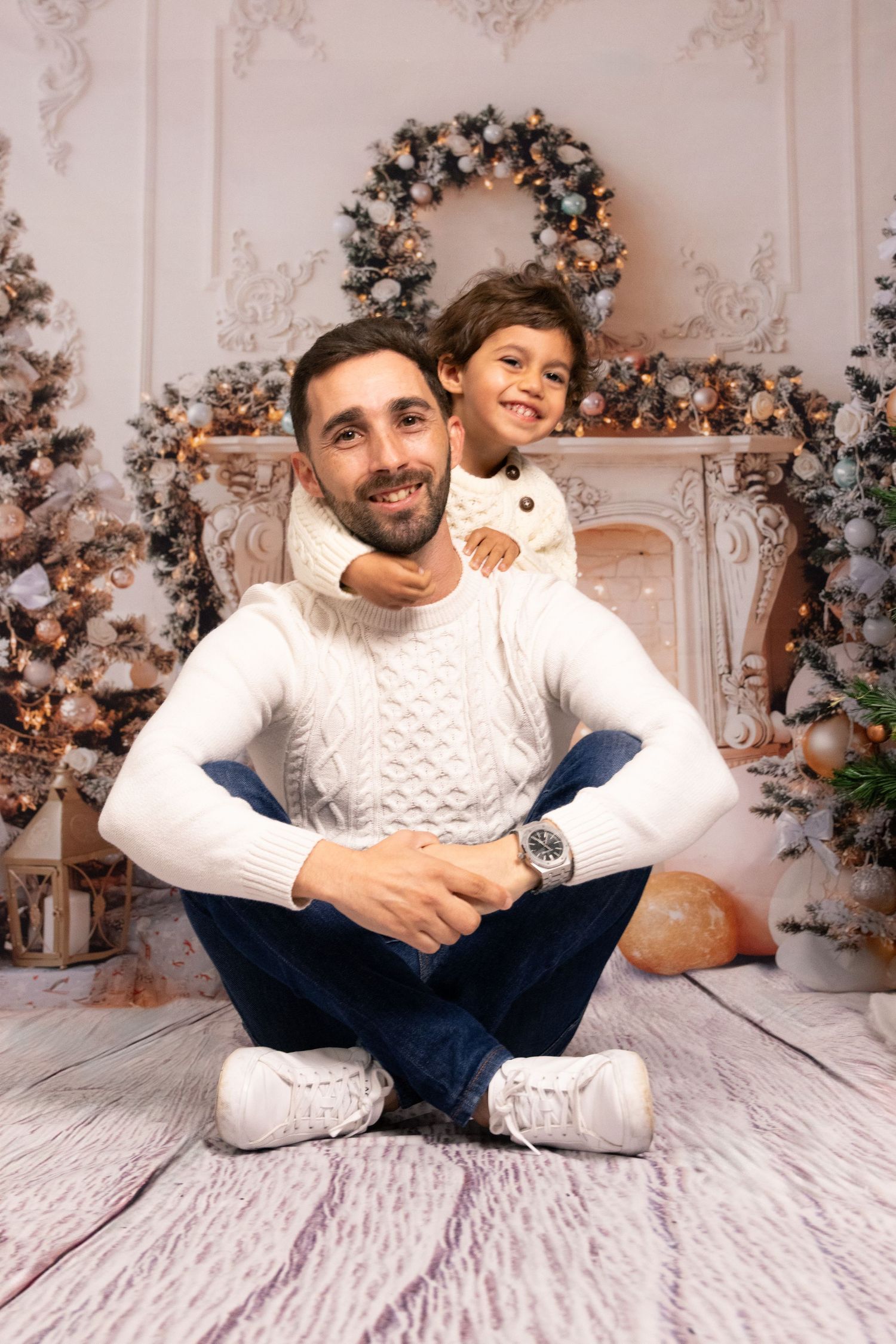 Person in white cable knit sweater and jeans poses playfully on white bedding with holiday decorations in background.