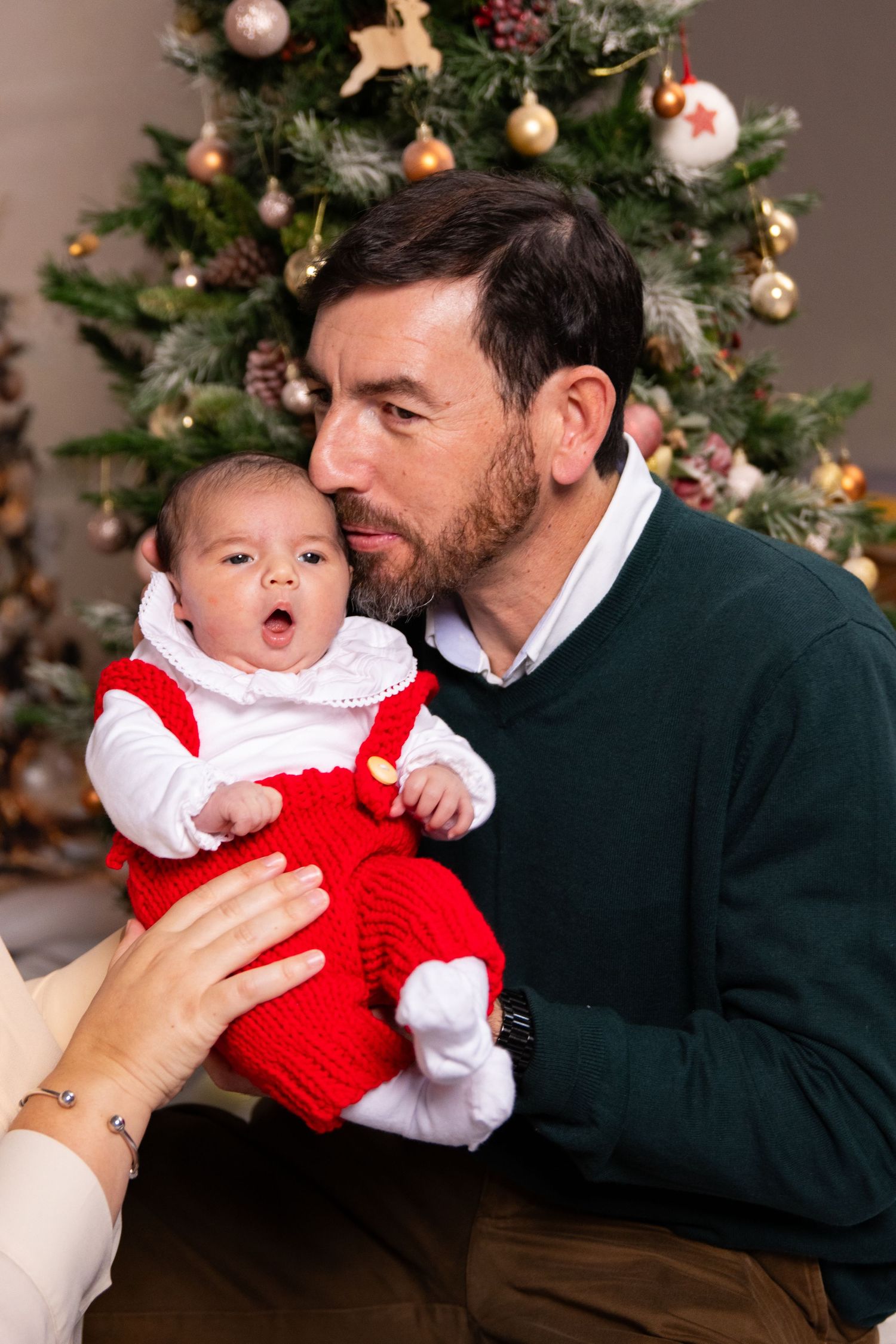 Baby in red holiday dress shares joyful moment with adult against Christmas tree backdrop.