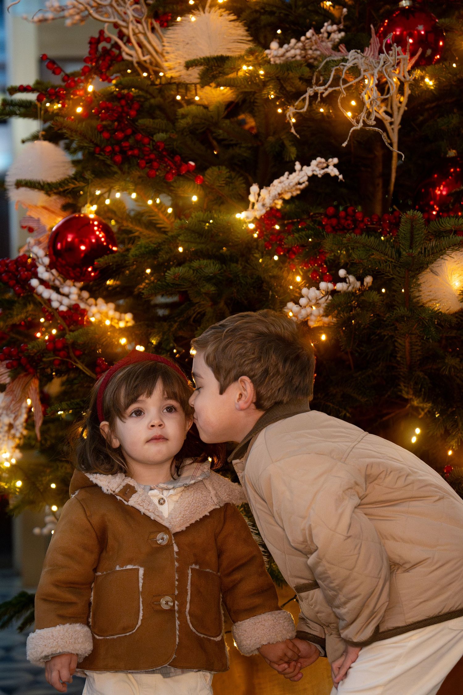 Two children in brown coats share a secret near decorated Christmas tree with glowing lights.
