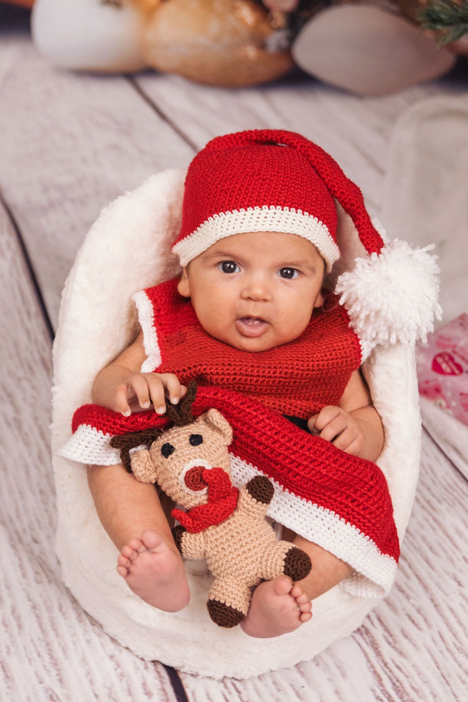 Baby wearing red knit Santa hat lies in basket with plush monkey toy on white wooden floor.