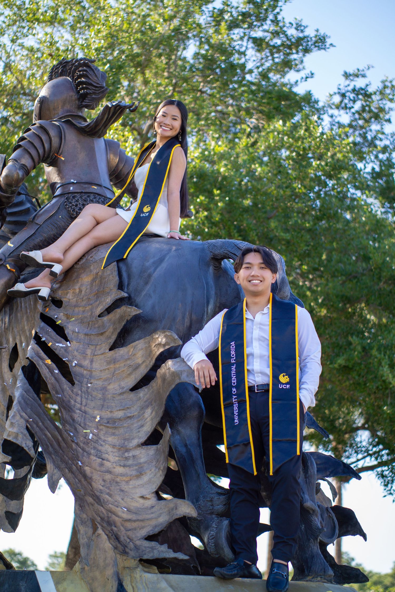 Graduates posing on a bronze statue wearing academic stoles and regalia during commencement celebrations.