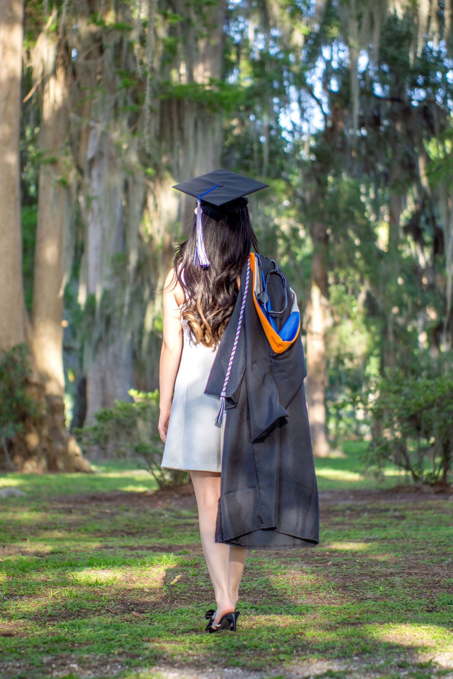 Graduate in black gown and cap walking away on a path through trees.