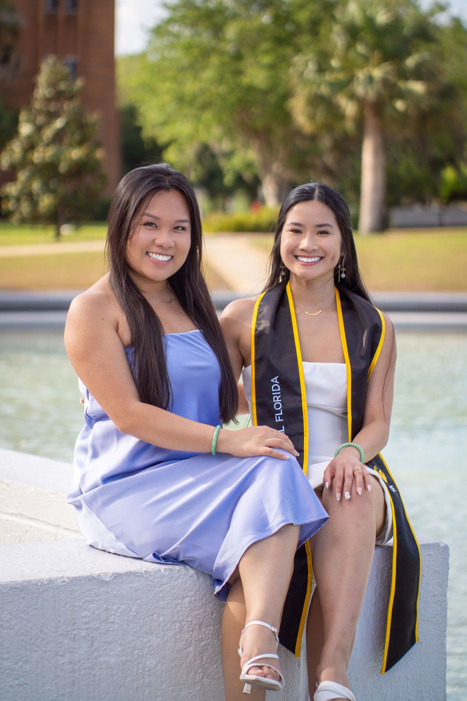 Two graduates in purple and white outfits sitting by a fountain during graduation celebrations.