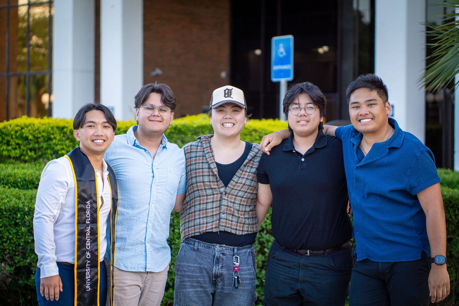 Group photo of graduates in casual attire and graduation stoles outside a campus building.