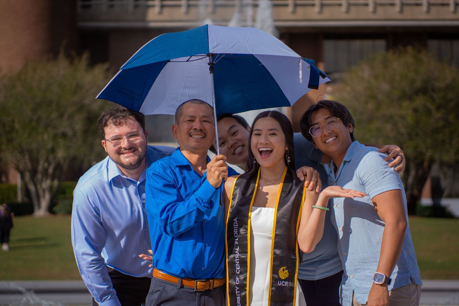 Graduates sharing a happy moment under a blue and white umbrella during commencement celebrations.