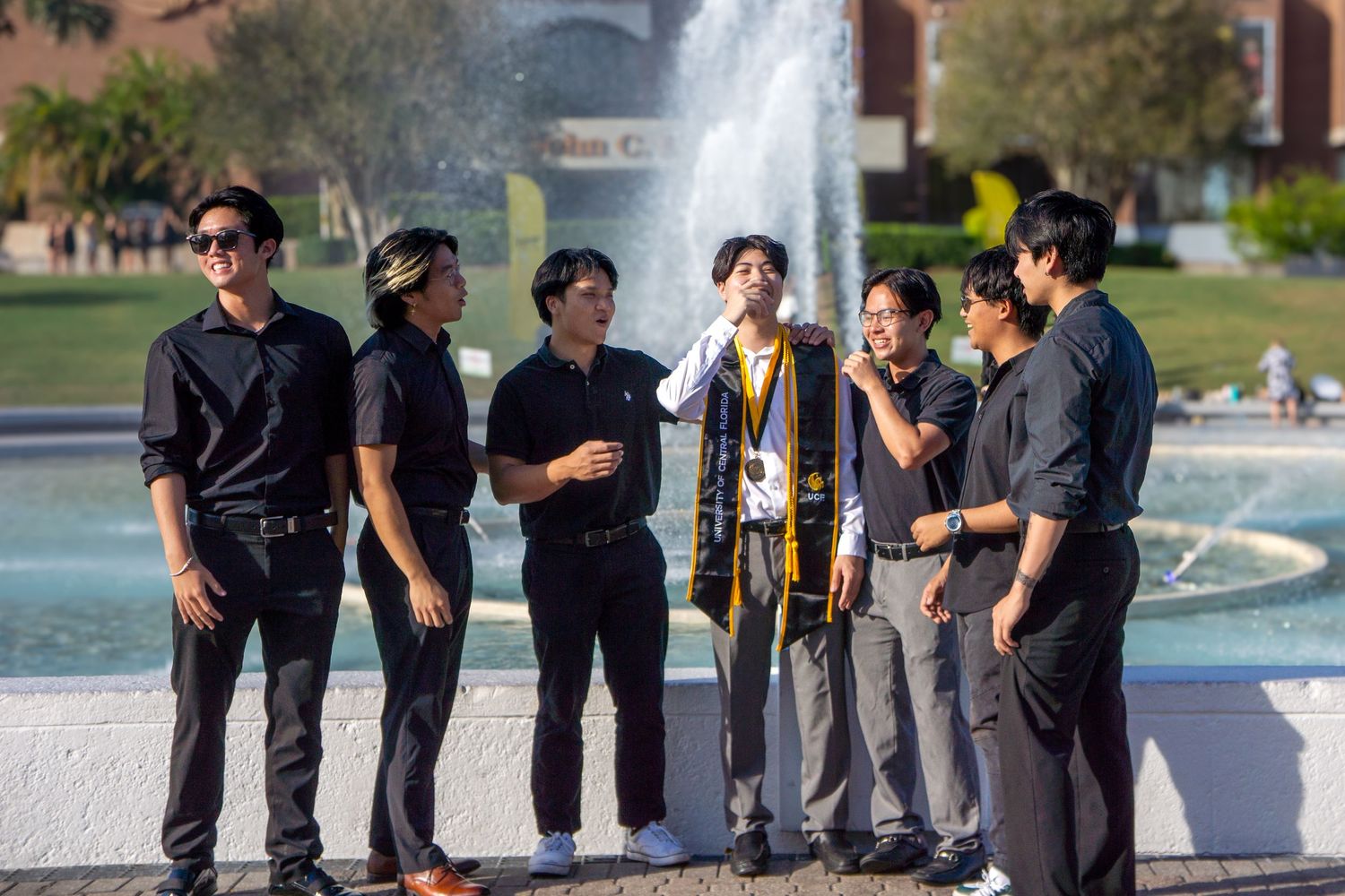Group of friends gathered near a fountain on a sunny campus day wearing casual attire.