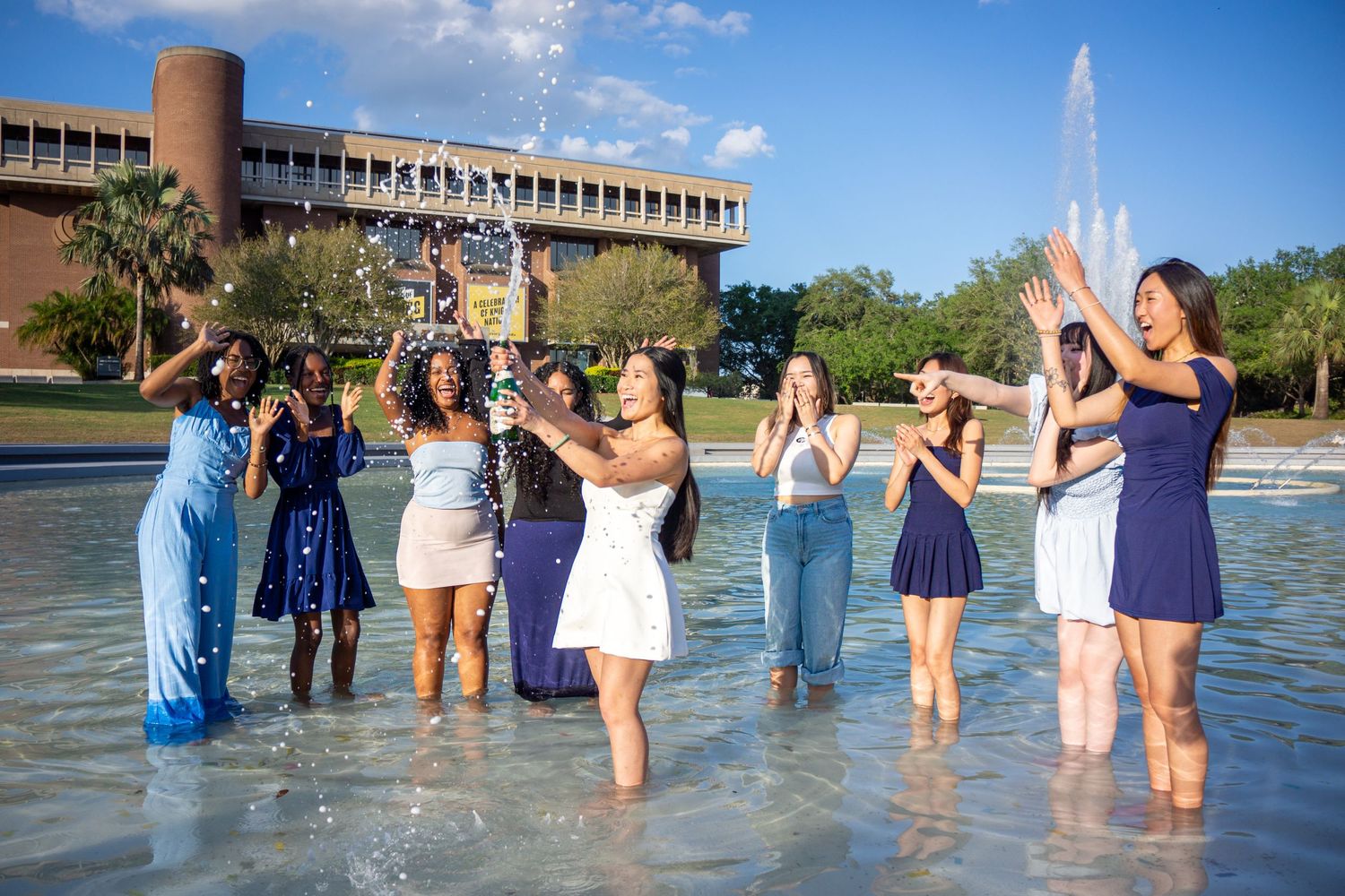 Joyful graduates celebrating by a fountain on their graduation day, wearing a mix of blue, purple and white dresses.