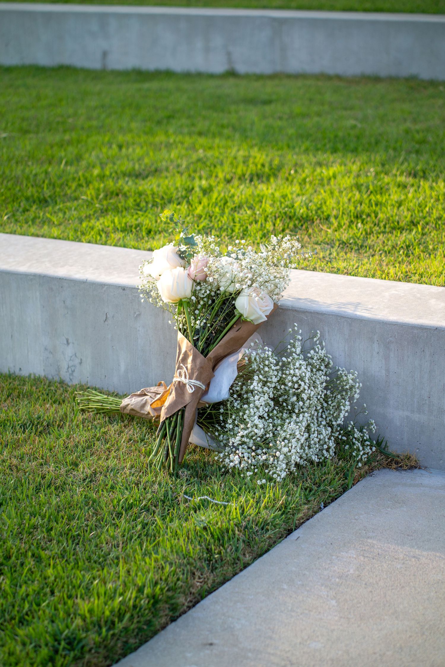 White floral bouquet and baby's breath lying on concrete ledge with green grass background.