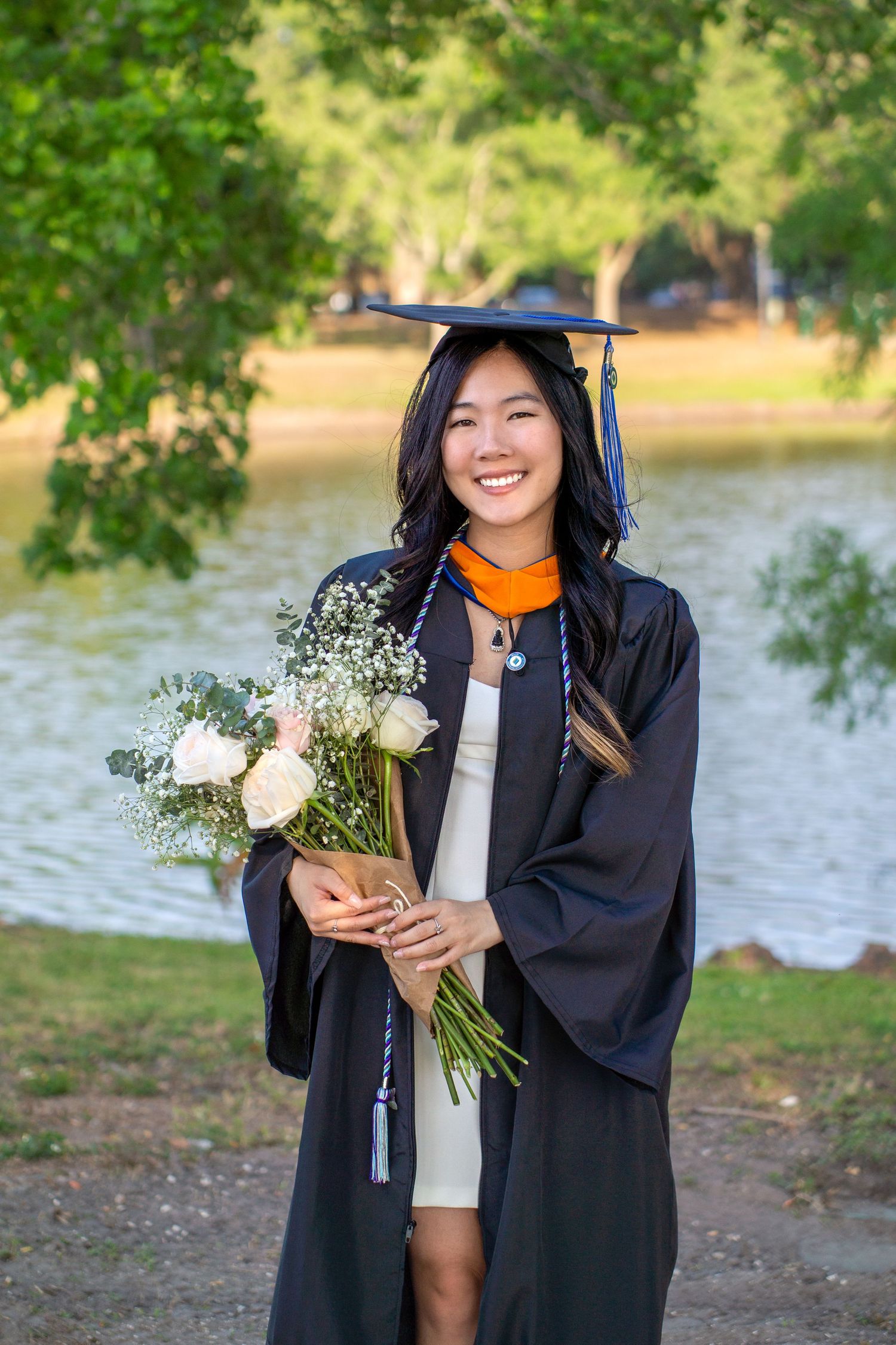 Graduate in black cap and gown holding a bouquet of white flowers posing by a lake.