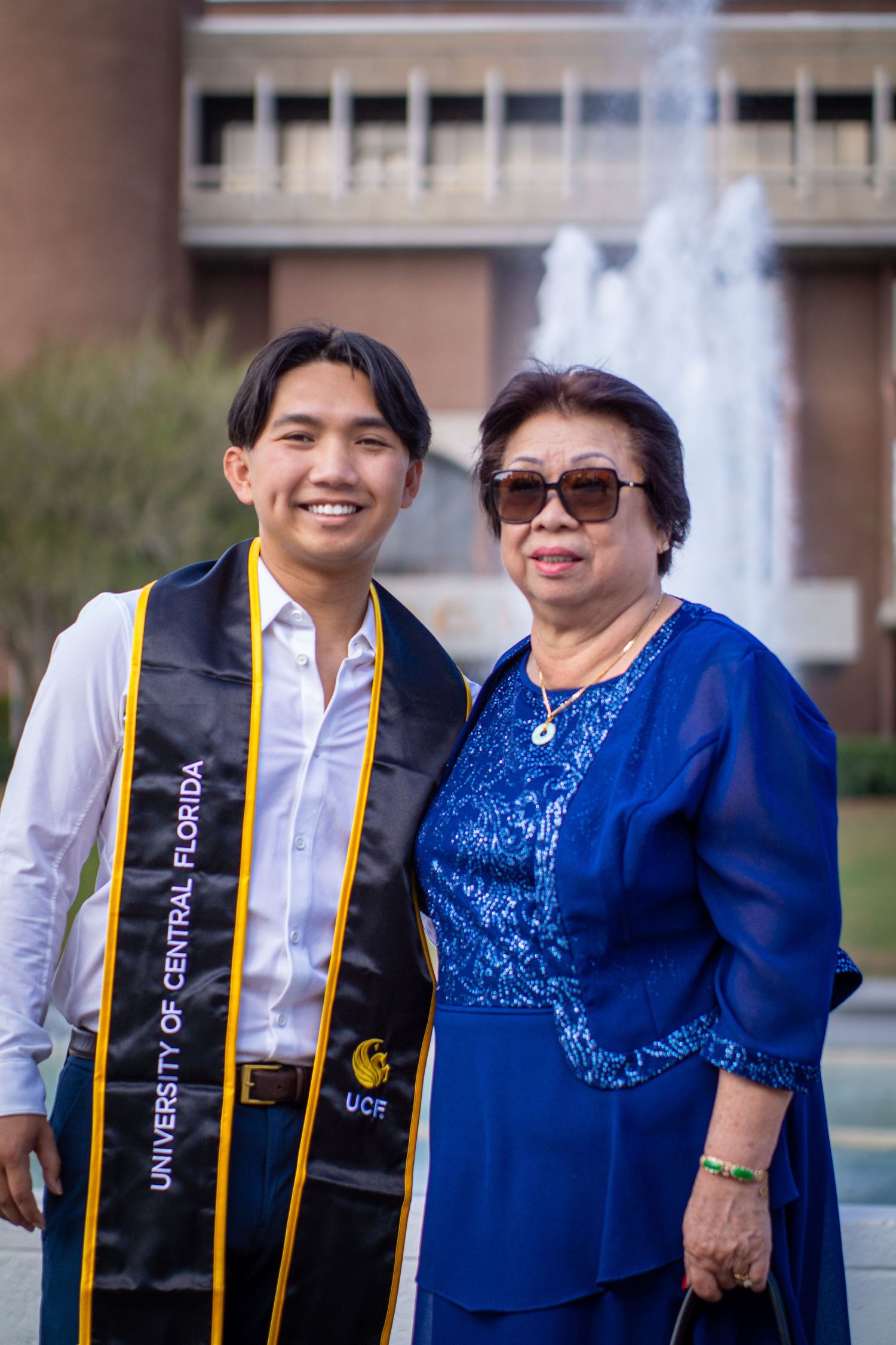 Graduate in academic regalia and guest pose in front of campus building with fountain.