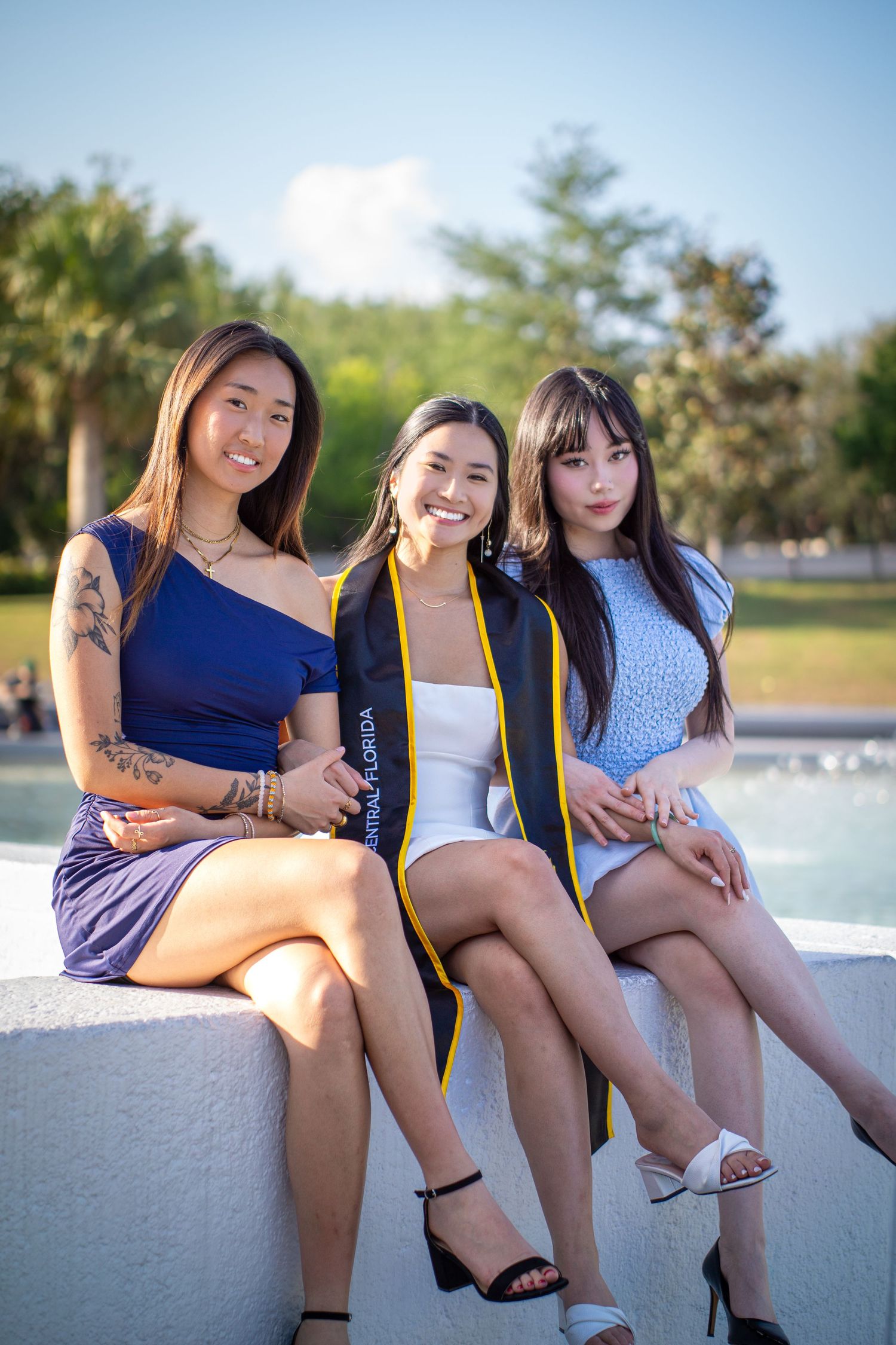 Three graduates in formal attire sitting by fountain on sunny day.