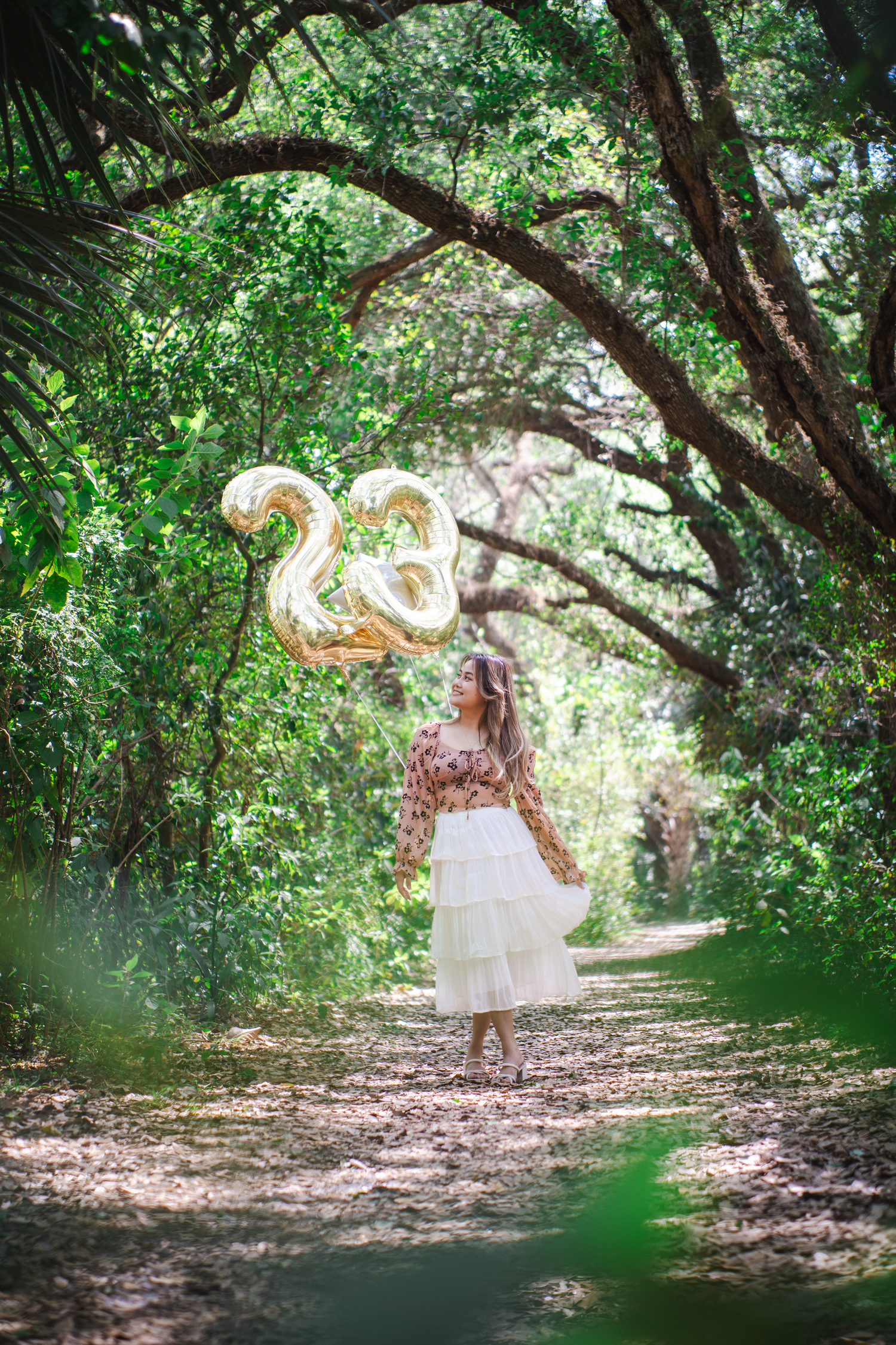 Gold number balloons under a natural forest archway on a sunlit path.