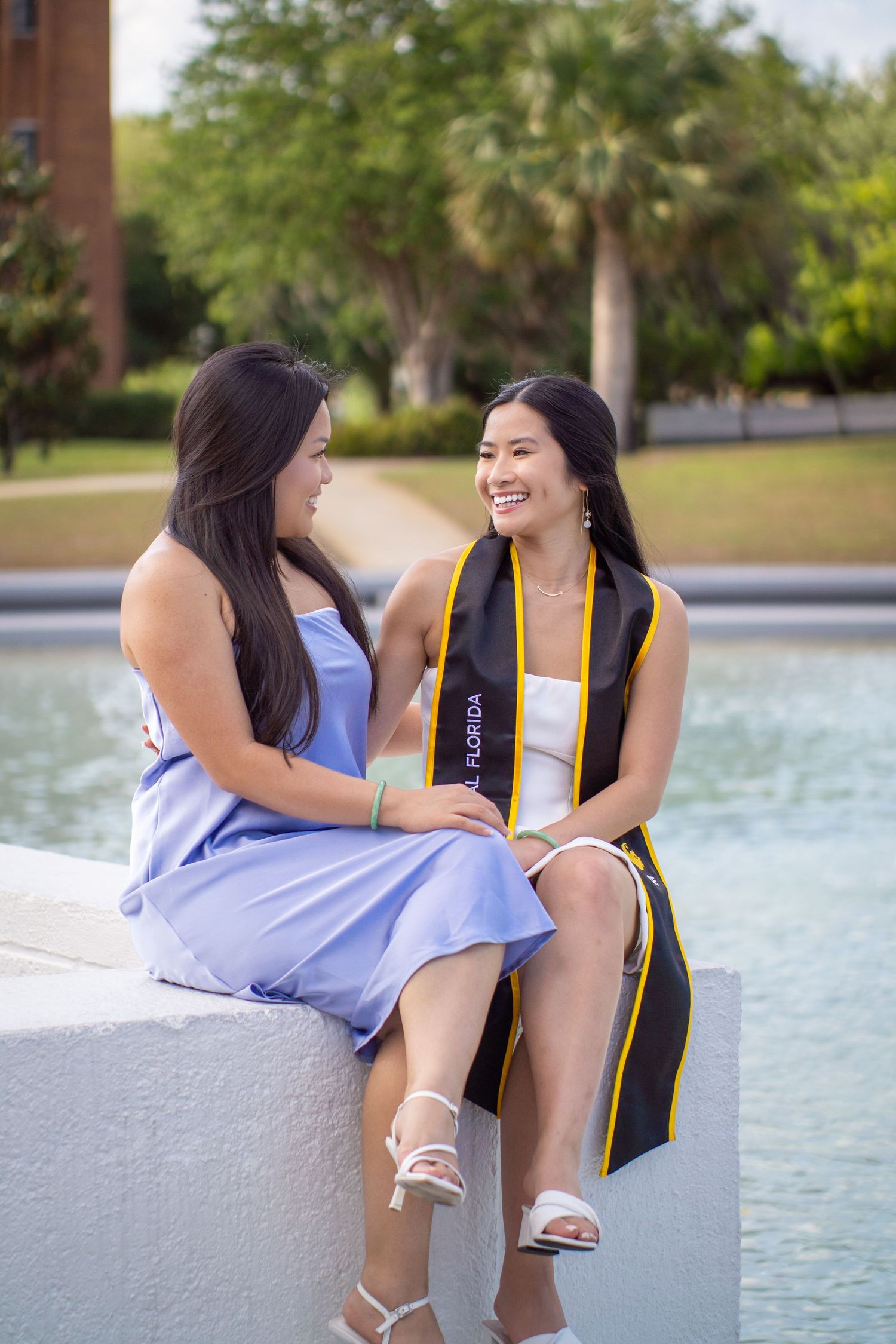 Two graduates in black and purple attire sitting by a fountain on campus during graduation celebrations.