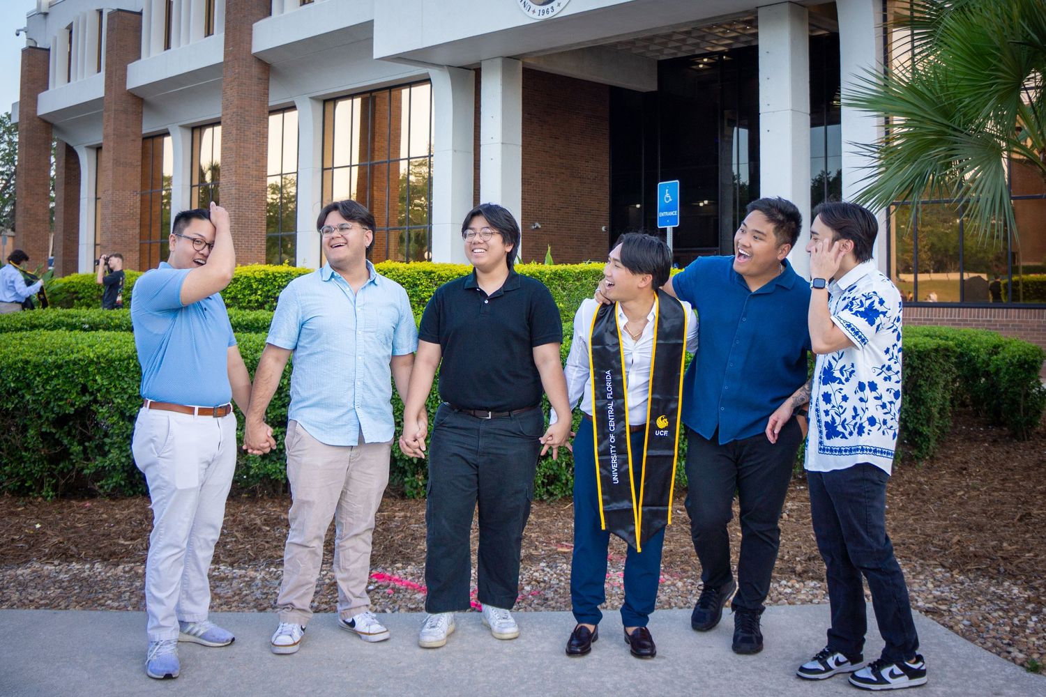 Group of graduates in casual attire sharing a lighthearted moment outside a building.