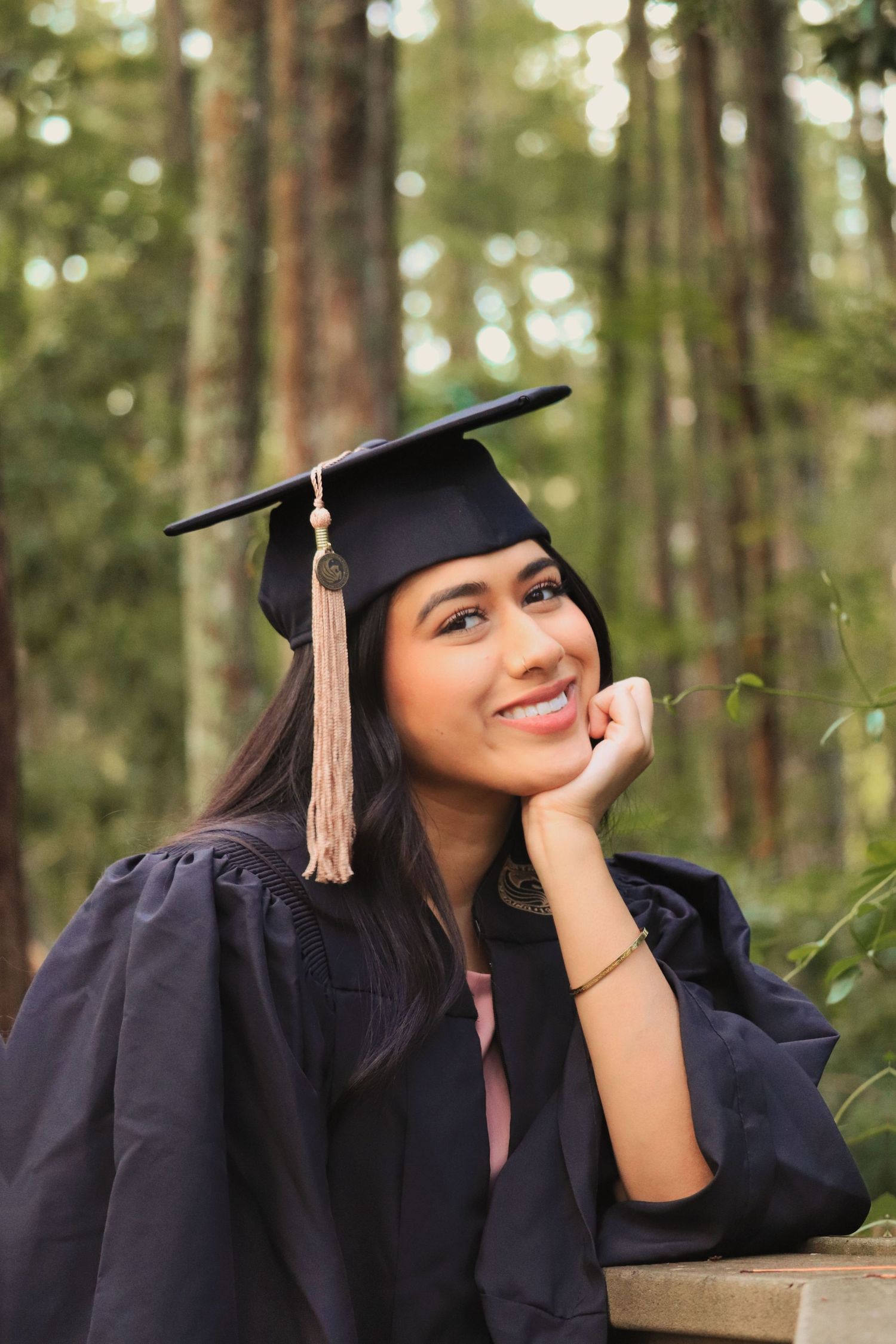 Graduate wearing traditional cap and gown poses thoughtfully in a peaceful forest setting.