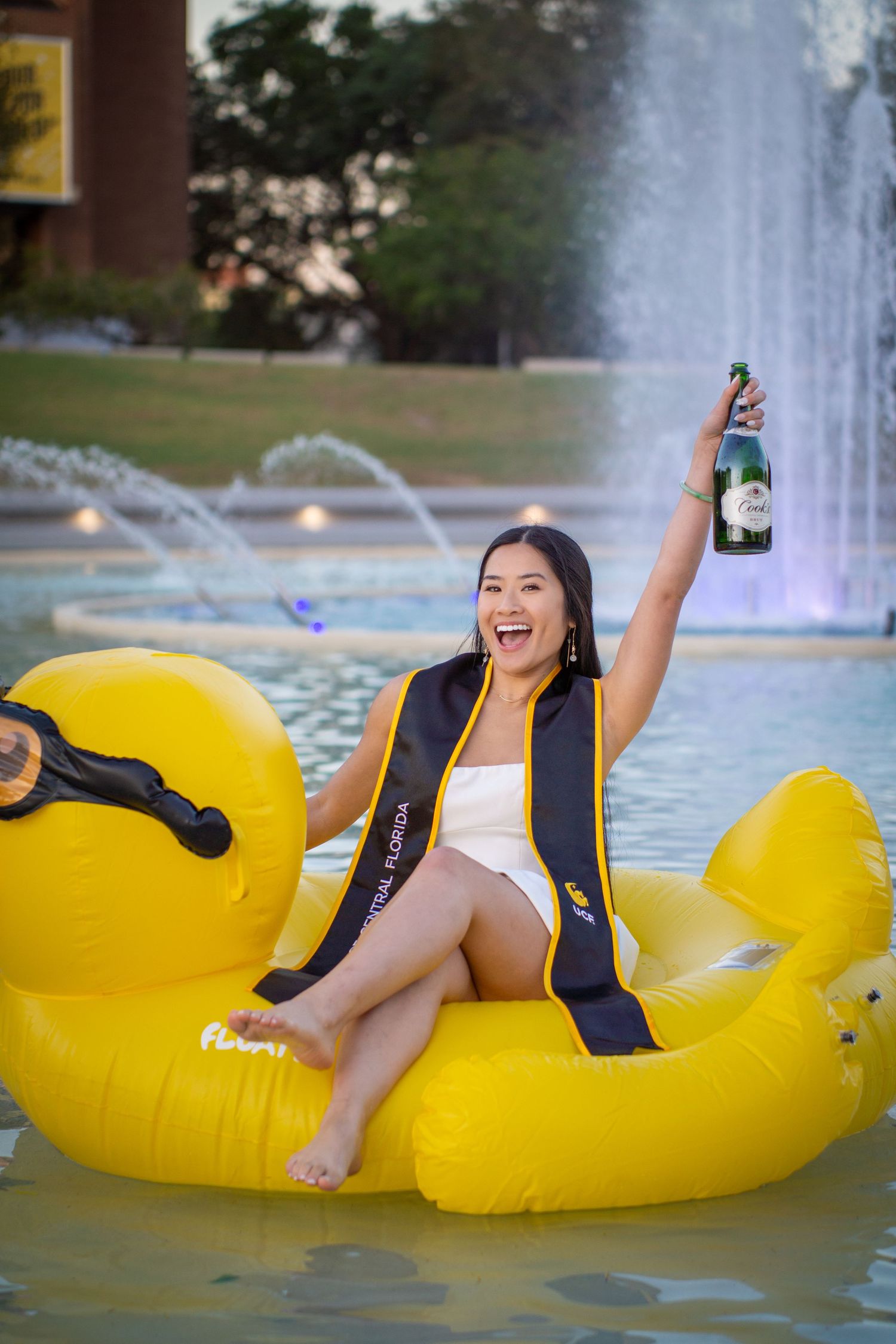 Graduate in academic regalia celebrates on a giant yellow rubber duck float in a fountain.