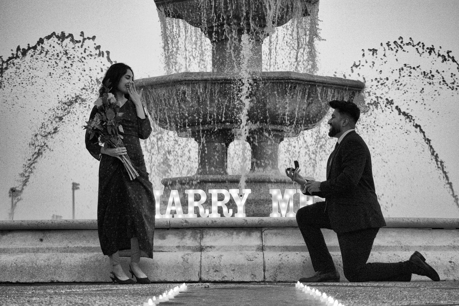 Black and white photo of proposal scene by fountain with 'Marry M' sign.