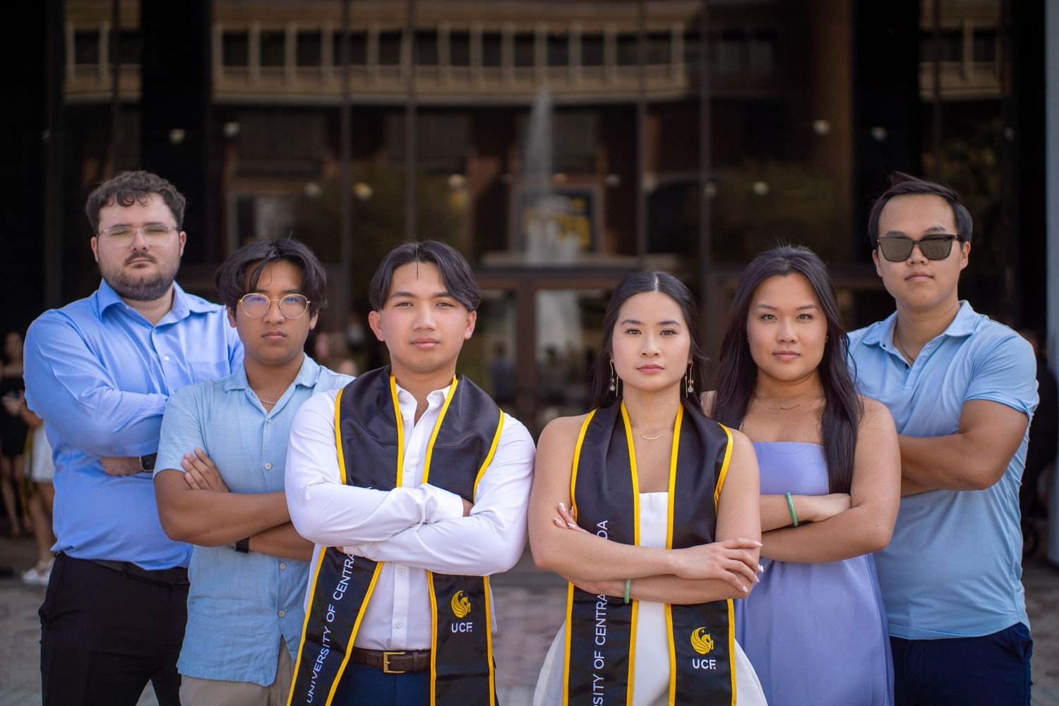 College graduates in formal attire and graduation stoles posing confidently in front of a building.