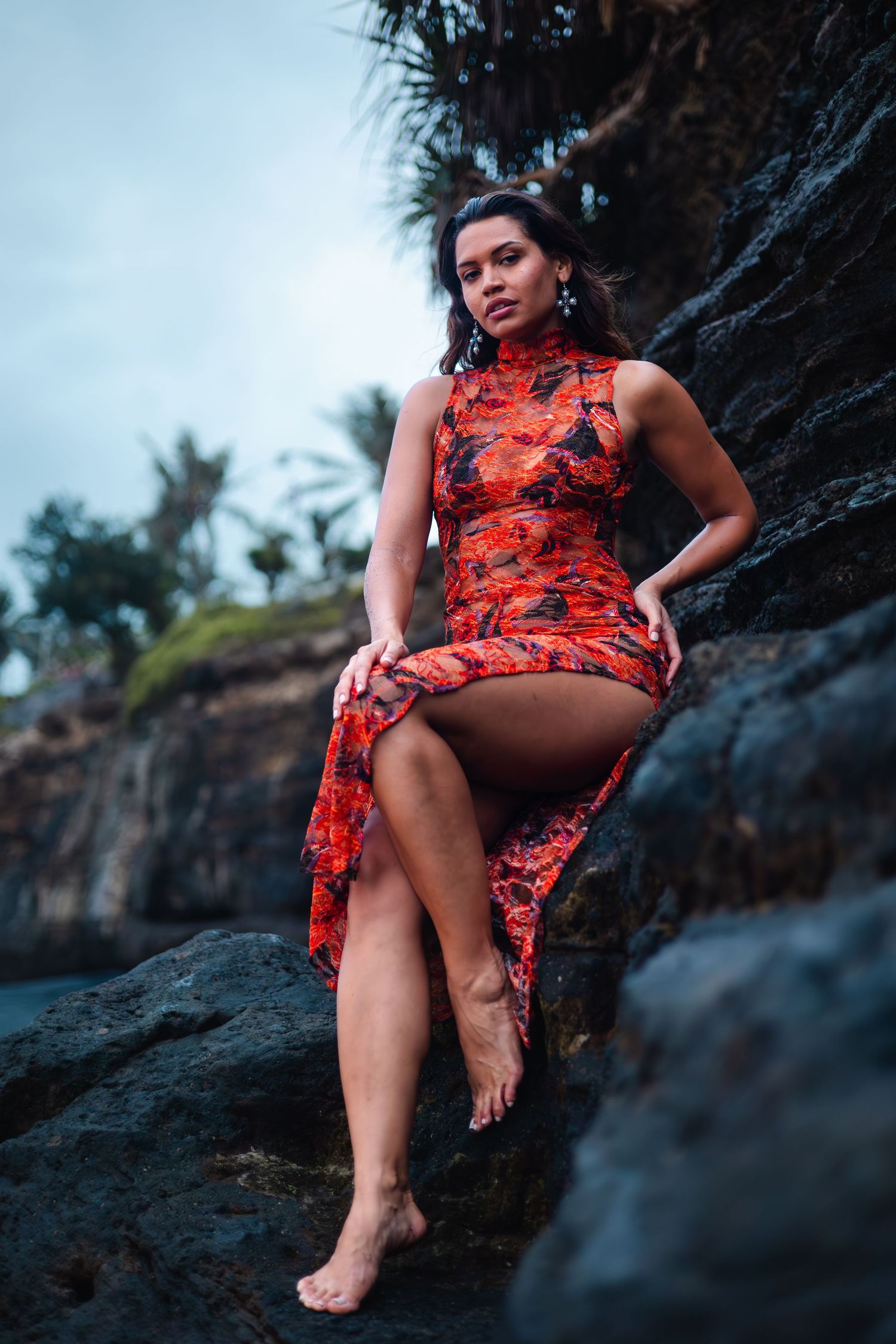Dramatic portrait in red Chinese-style dress against rocky coastal backdrop.