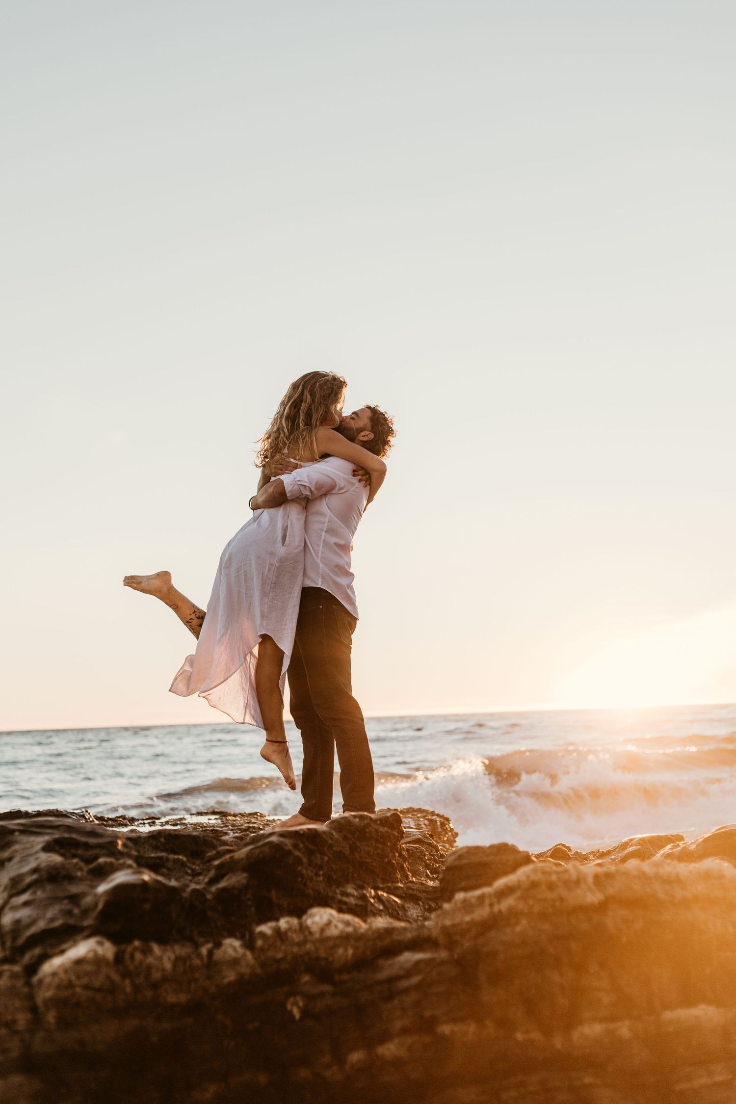 A romantic couple embrace on coastal rocks during golden hour with ocean waves crashing behind them.