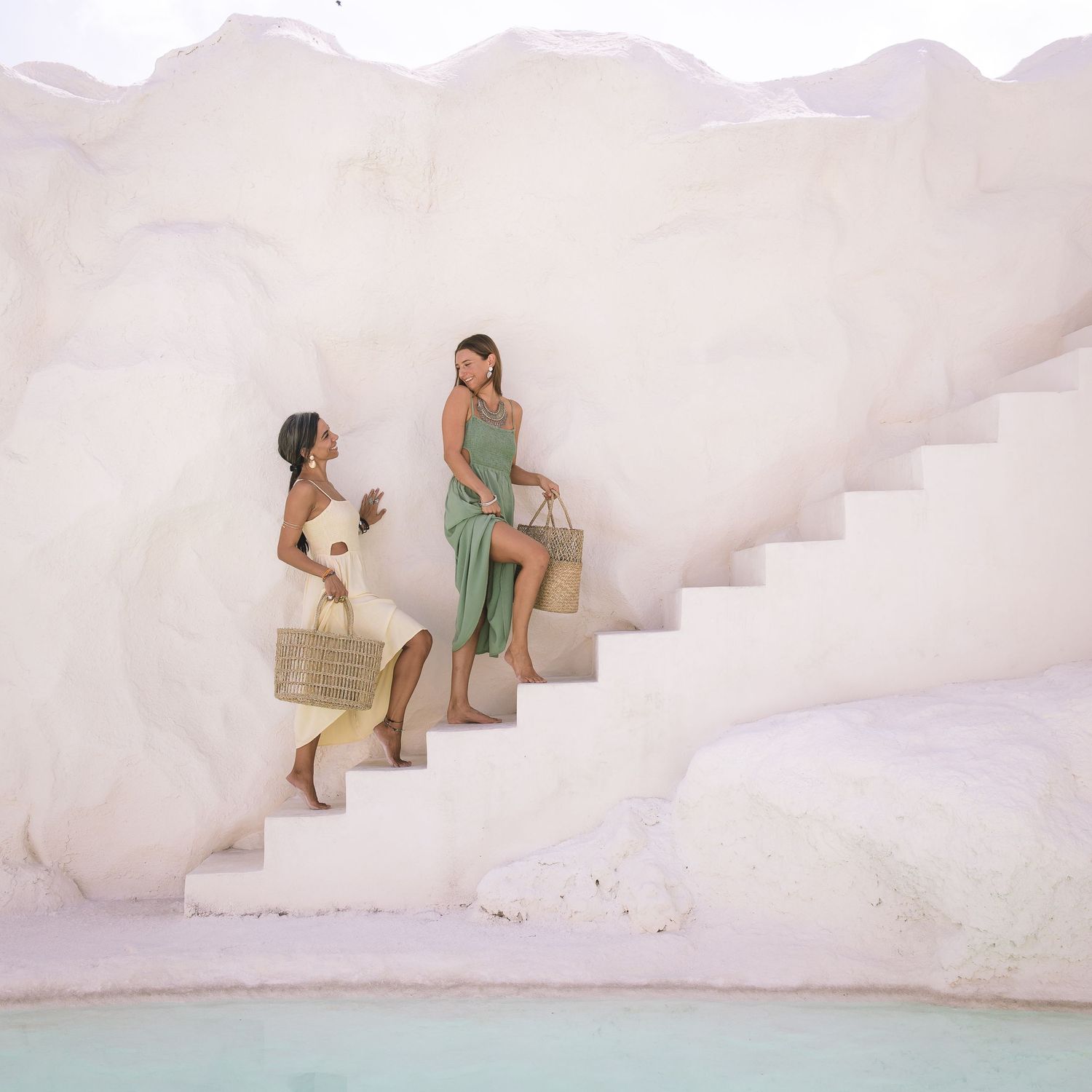 Two women in summer dresses sitting on white concrete steps overlooking a pool in a minimalist setting.