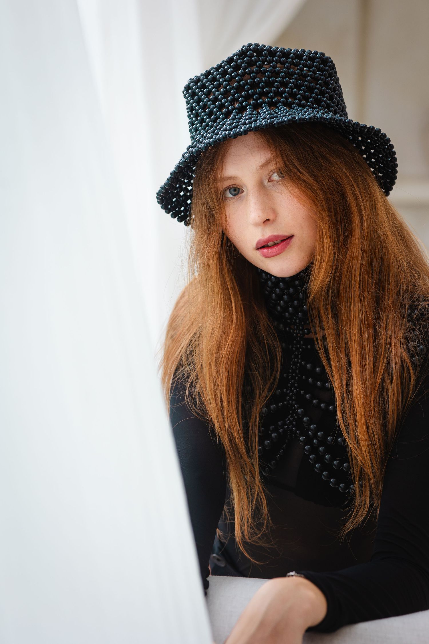Portrait of a person wearing a sparkly black hat and dark sweater with long red hair against a light background.