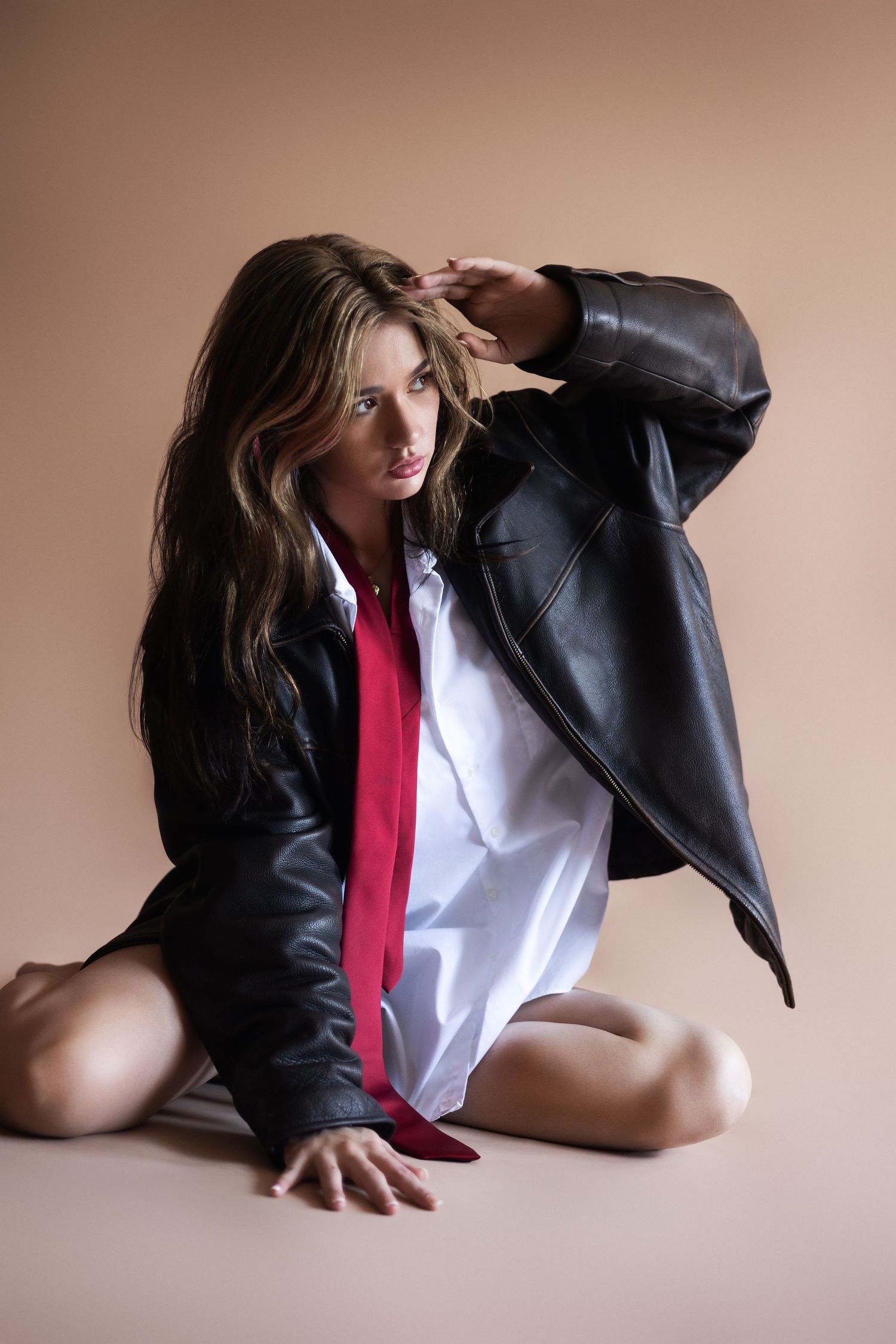 Model in an oversized black leather jacket and red tie poses seated against beige background for fashion photoshoot.