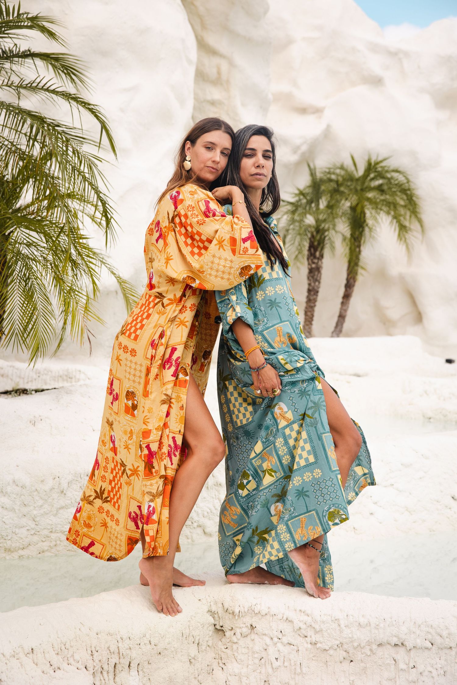 Two women in colorful bohemian maxi dresses pose against a palm tree and white sand tropical beach backdrop.