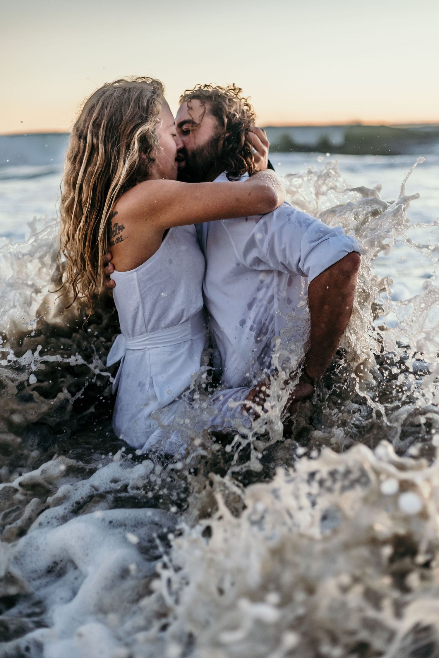 A couple in white clothing shares a passionate kiss as ocean waves splash around them.
