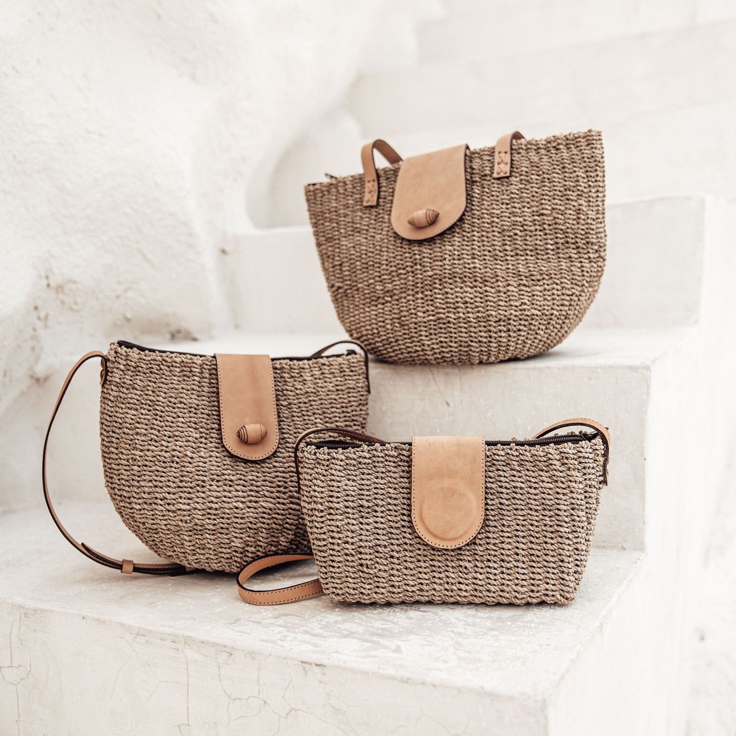 Three woven straw bags with leather accents displayed on white steps in ascending sizes.