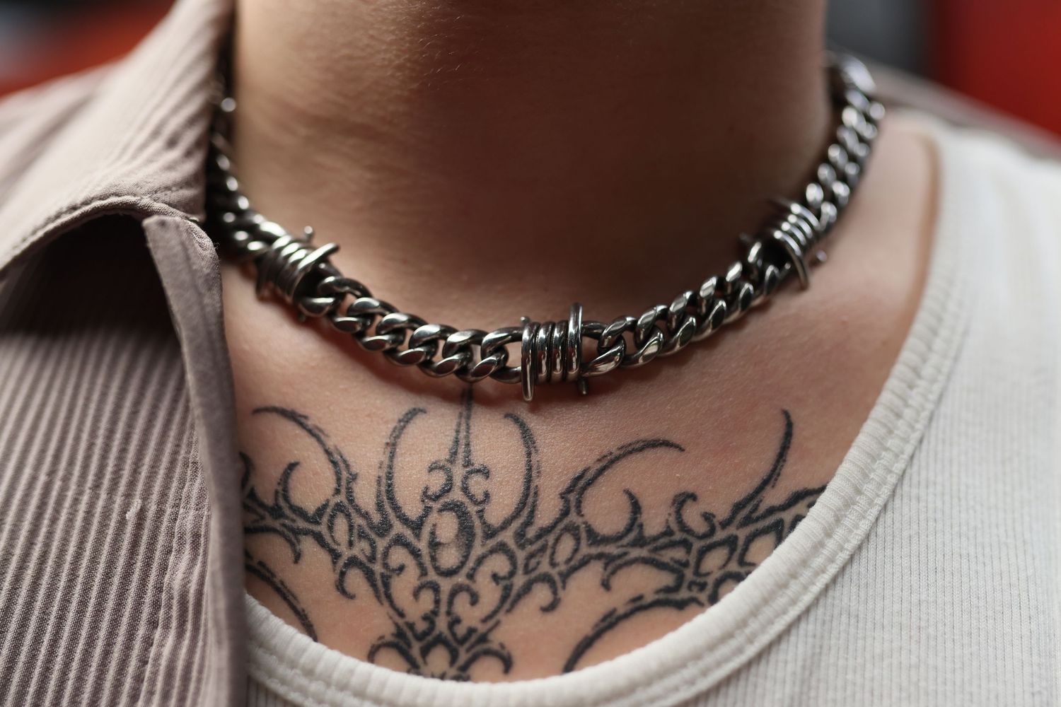 Close-up of a silver chain necklace and chest tattoo against a beige ribbed shirt collar.