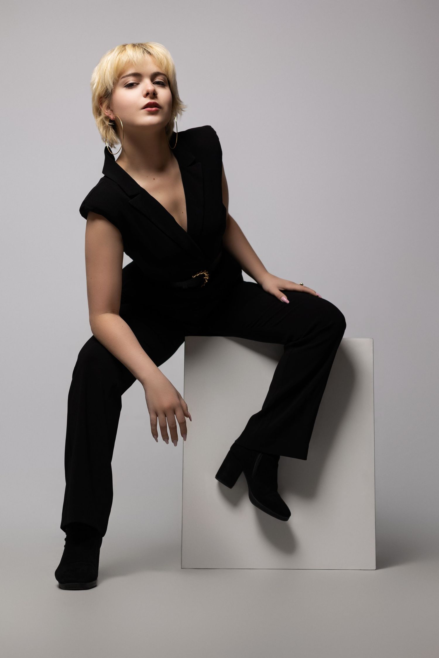 Stylish portrait of person in black velvet jumpsuit seated on white cube.