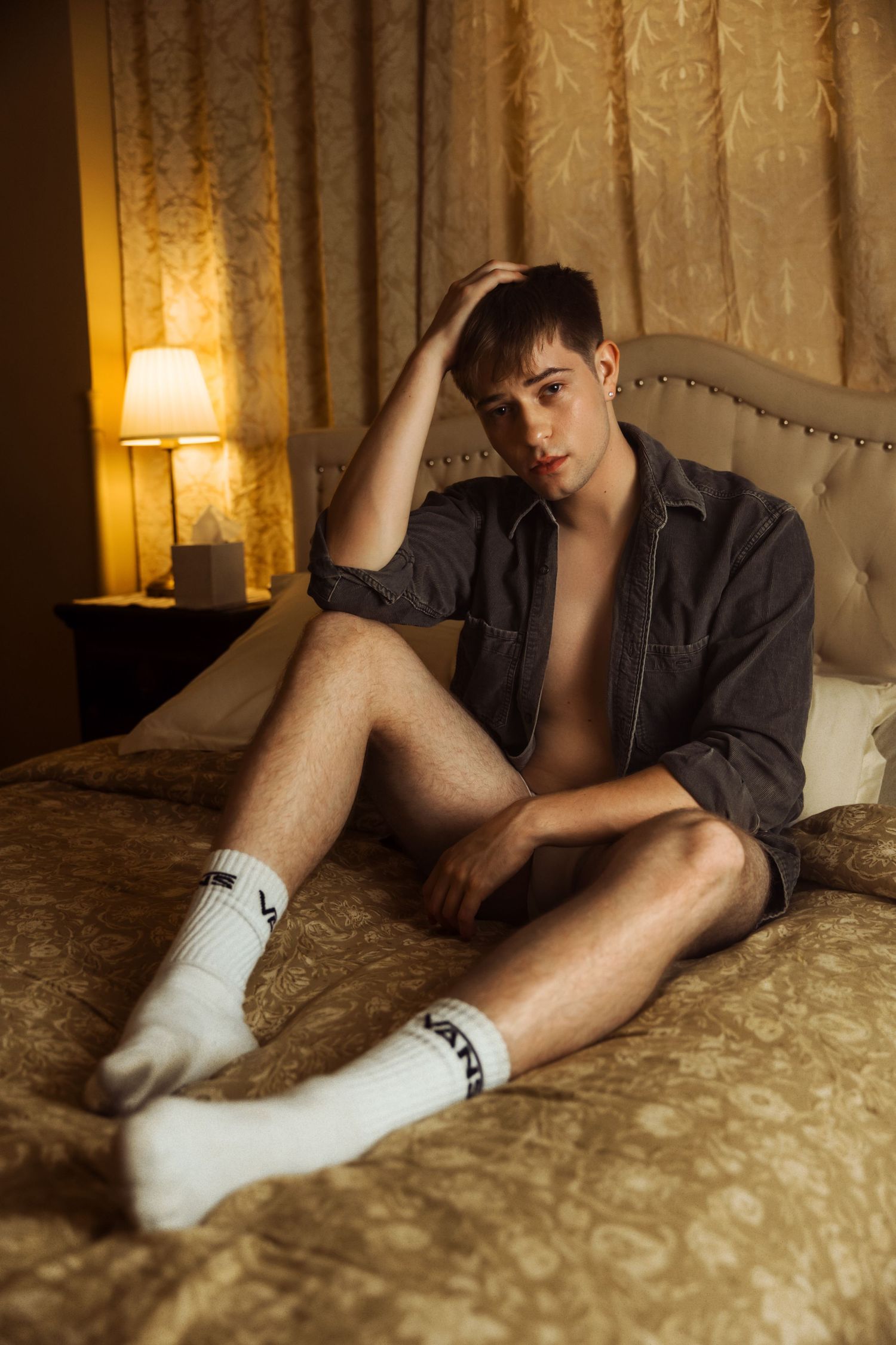Casual pose on bed wearing denim jacket and white socks with warm lighting.