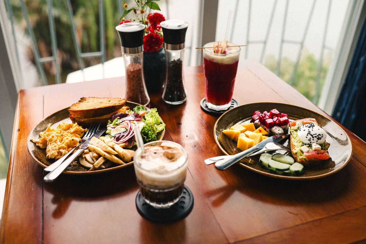 Two plates of casual dining food with side salads are served alongside iced coffee drinks on a wooden restaurant table.