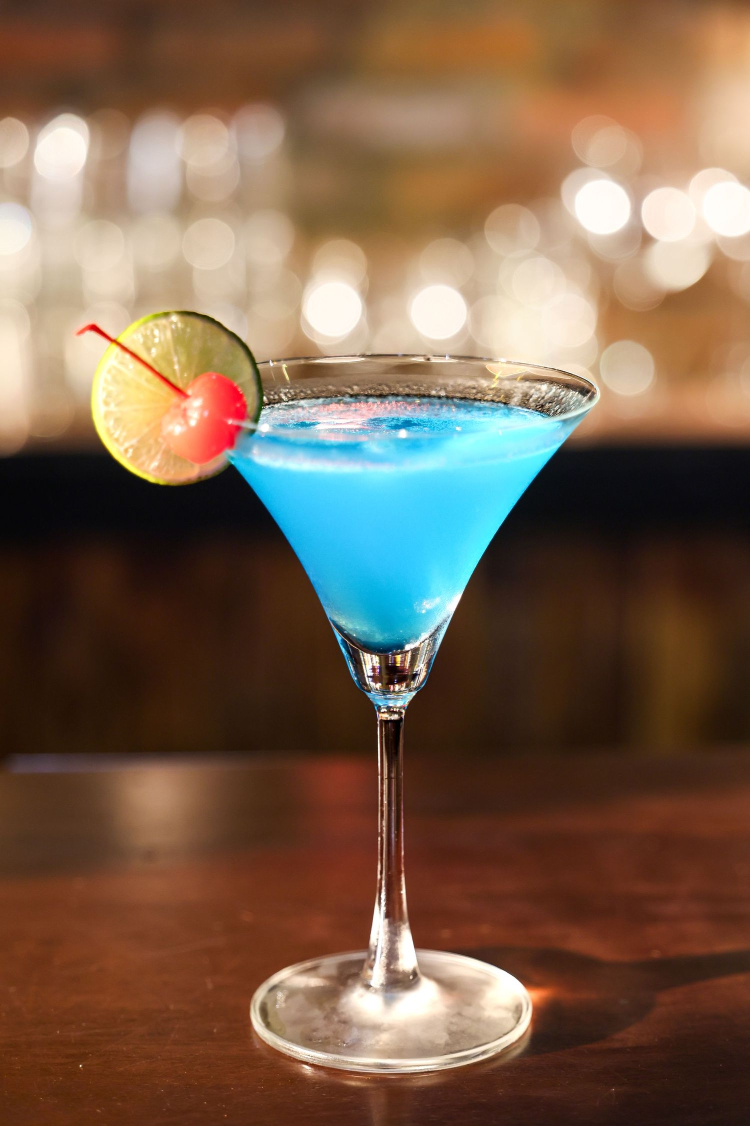 A bright blue martini cocktail in a clear glass garnished with a lime and cherry on a dark wooden bar counter.