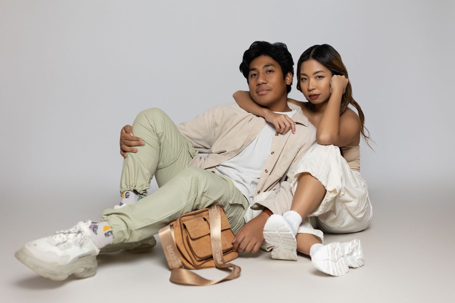 Two young models in casual white outfits sitting and lounging together on a light gray studio backdrop.