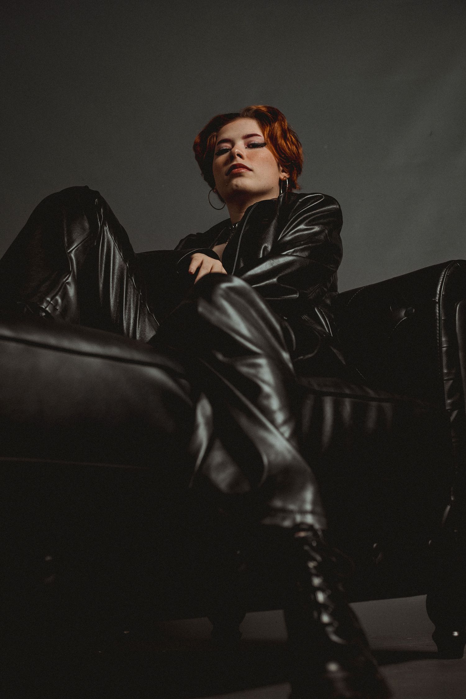 Moody portrait of person in all-black leather outfit reclining on dark couch.