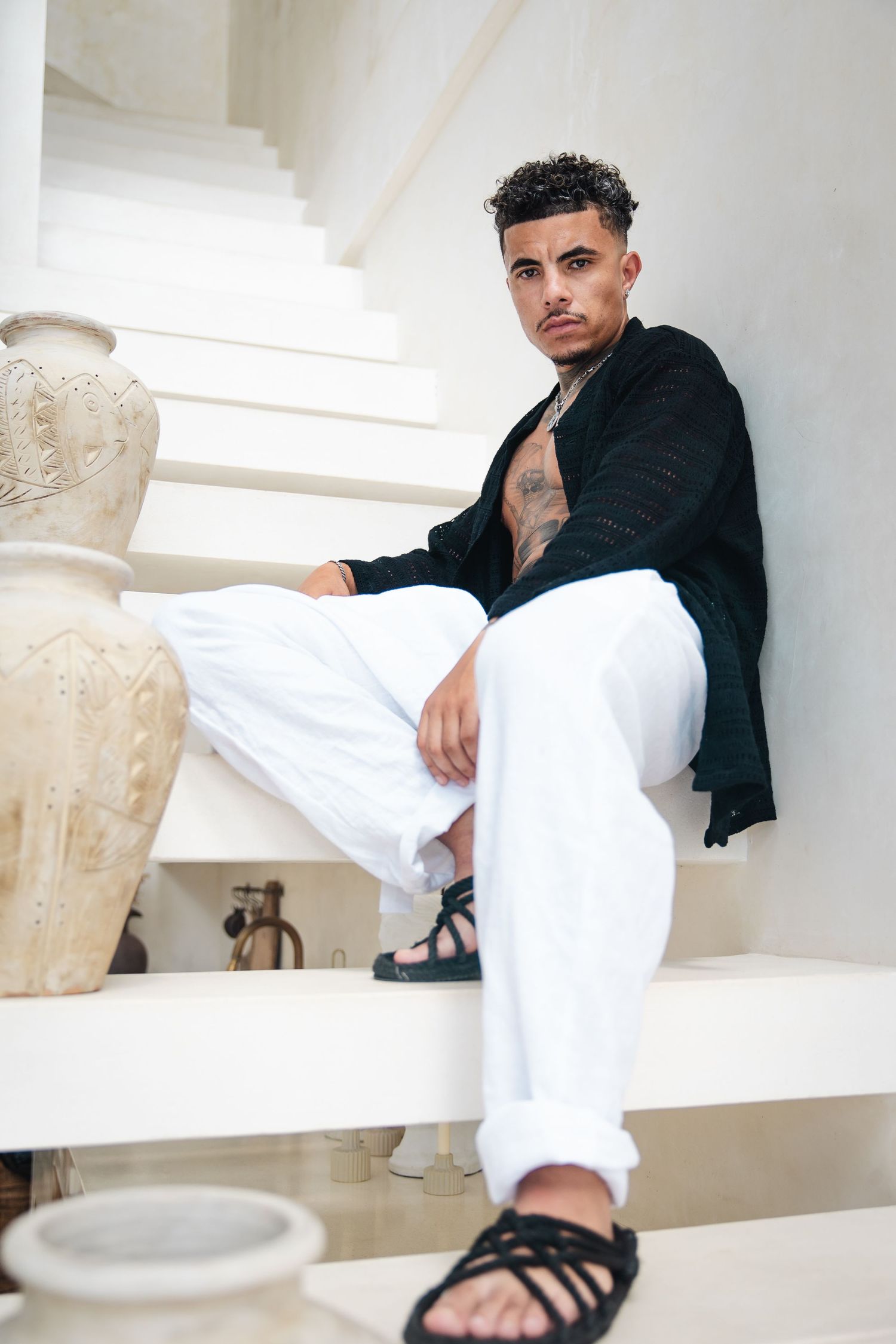 Trendy model in black shirt and white pants poses on white steps next to decorative vases wearing strappy sandals.