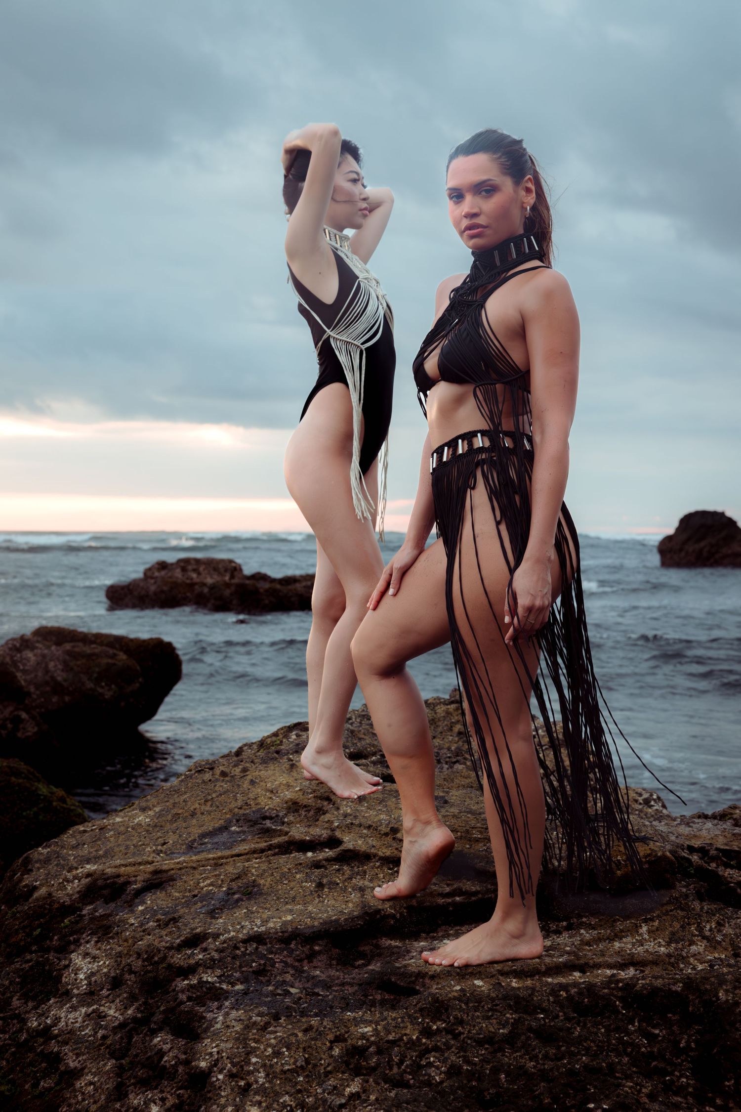 Two models pose dramatically on coastal rocks at sunset wearing stylized swimwear and fringe designs.
