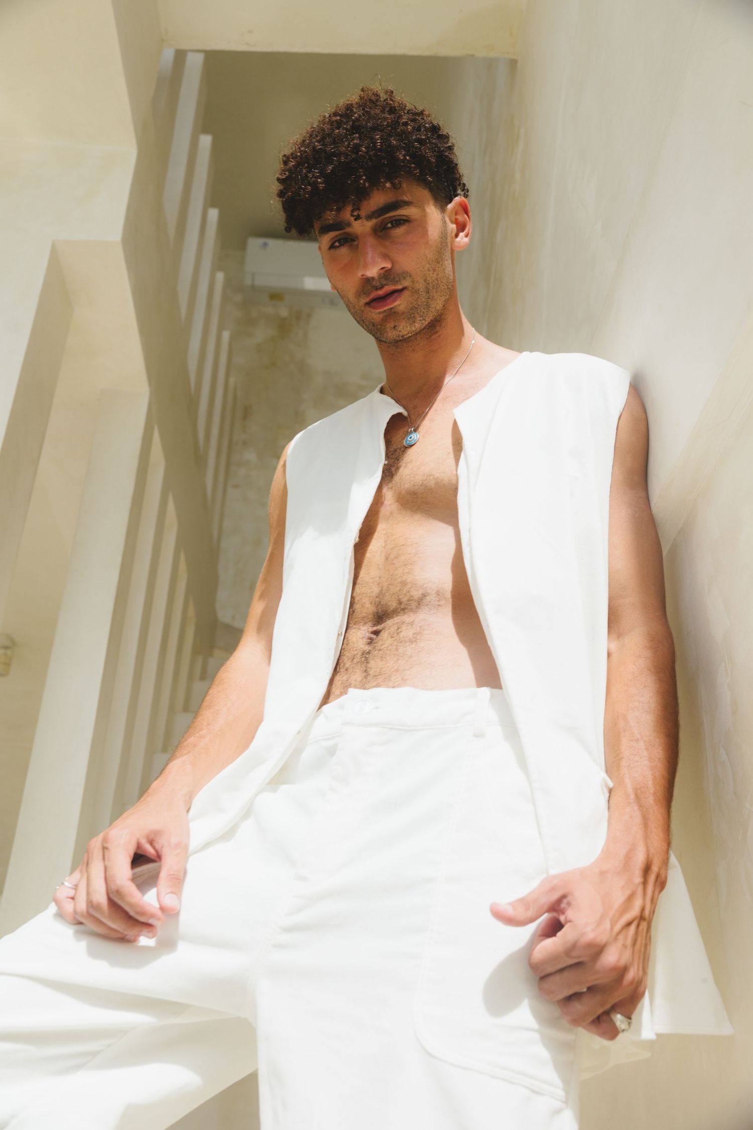 Fashion model wearing all-white ensemble with open vest against light wall background.