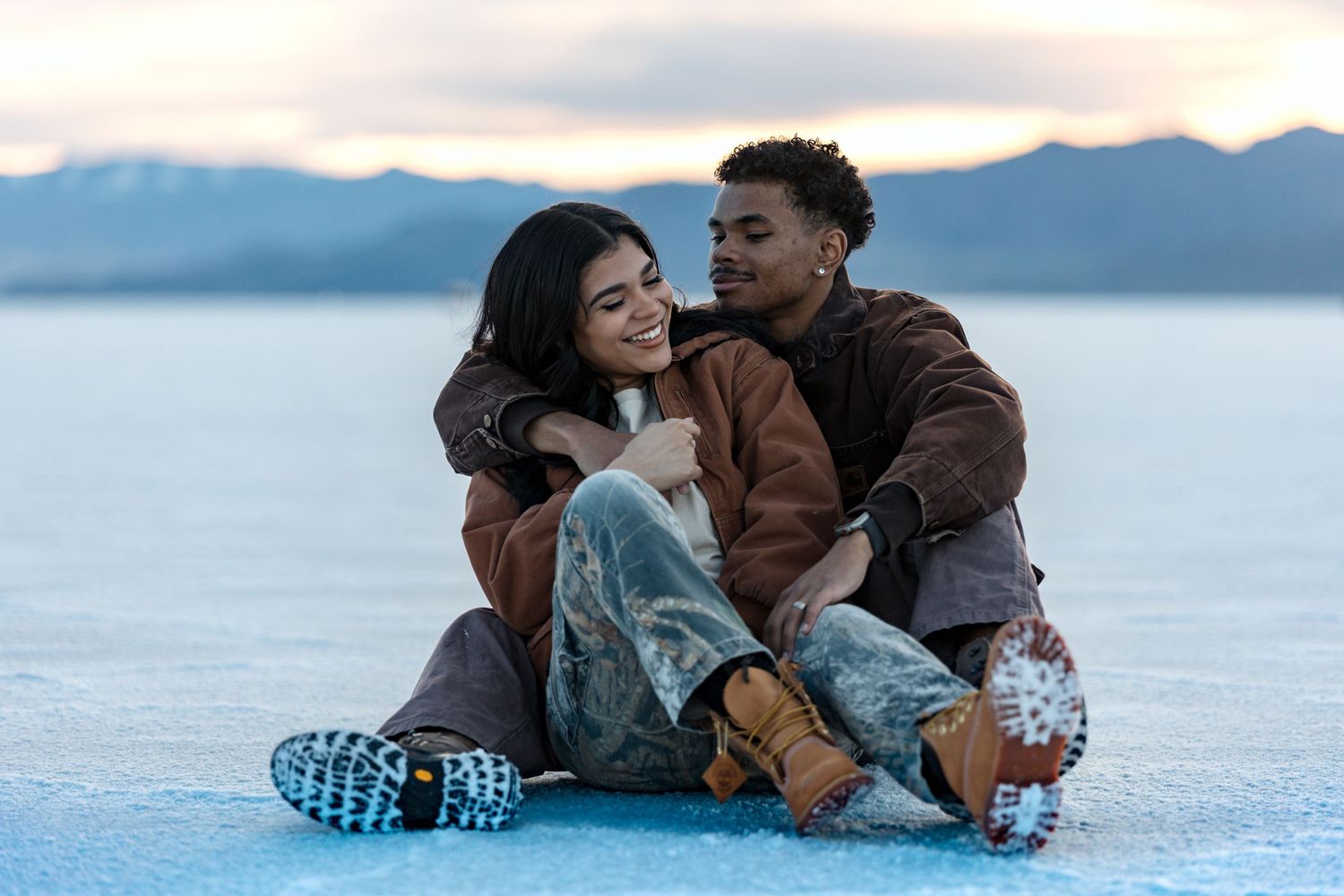 Couple sitting together on snowy ground with mountain backdrop.