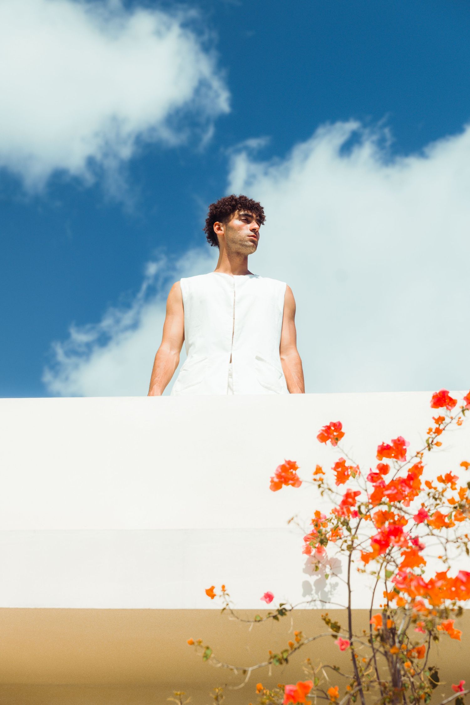 A figure in a sleeveless white outfit stands against a bright blue sky with orange flowers blooming in the foreground.