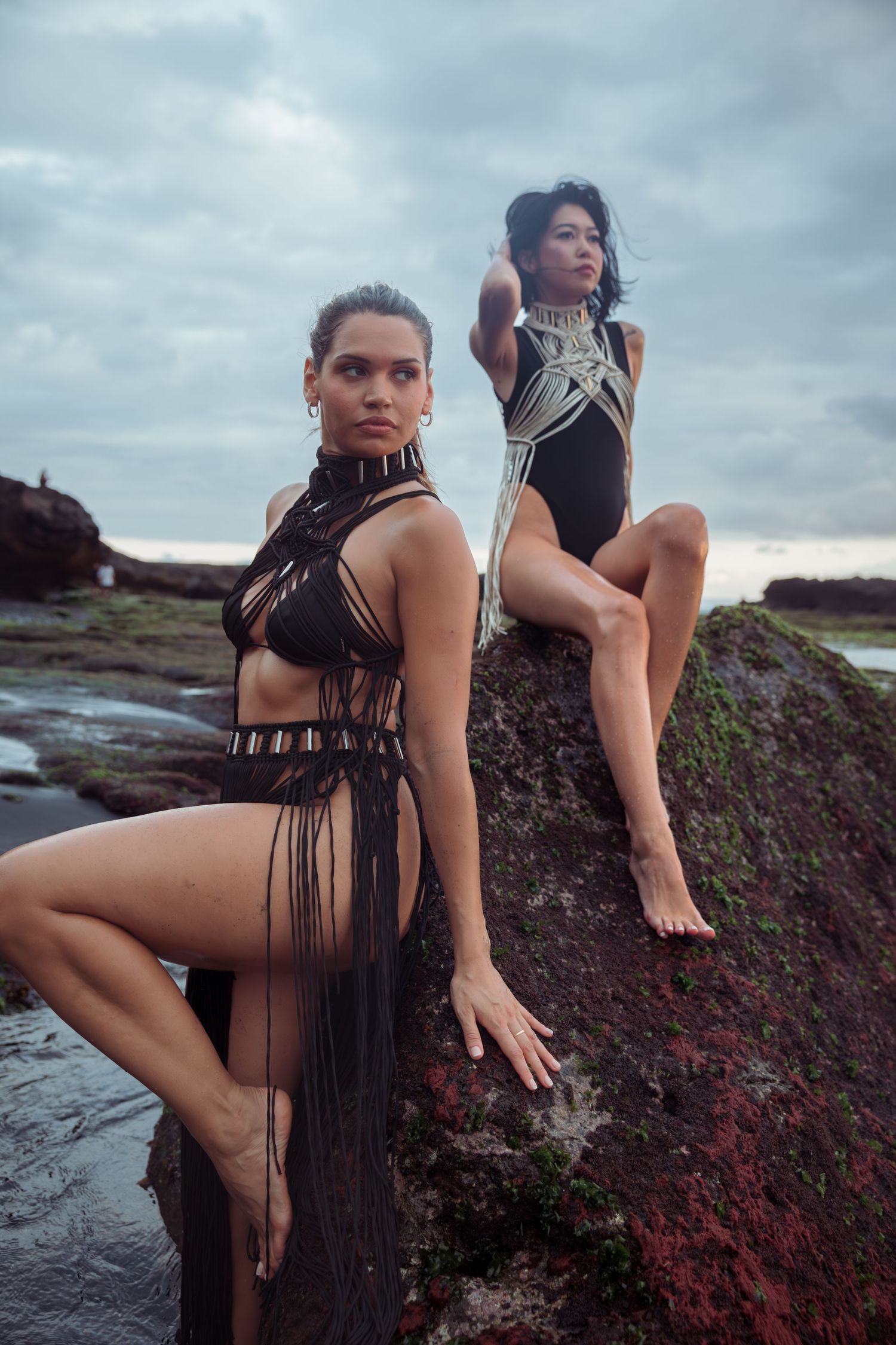 Two models in dark swimwear pose dramatically on volcanic rocks near the ocean under cloudy skies.