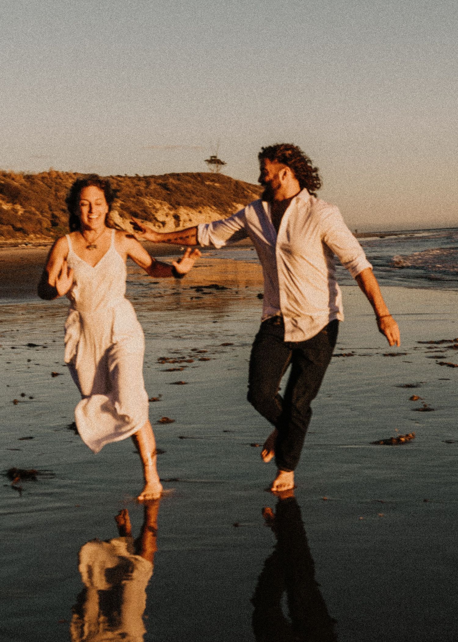 A couple runs playfully along a beach at sunset with their reflections in the wet sand.