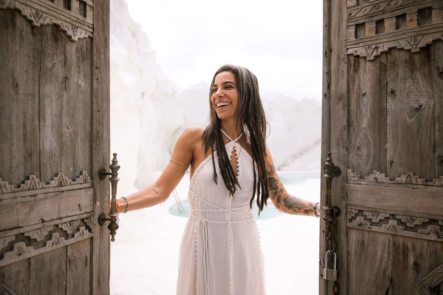 A person in a flowing white dress stands between ornate wooden double doors with a joyful expression.