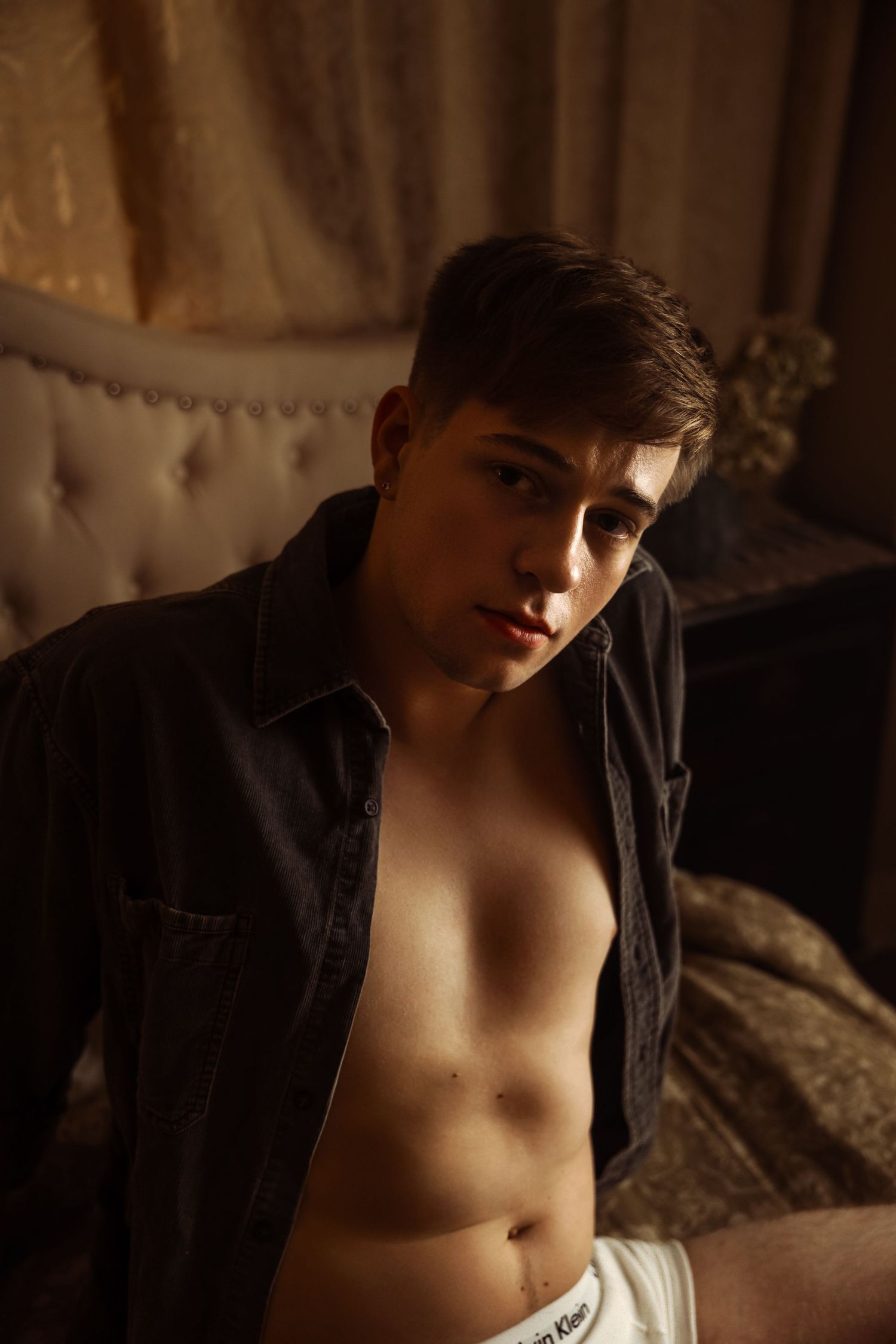 Person in unbuttoned dark shirt sitting on bed in dramatic lighting.