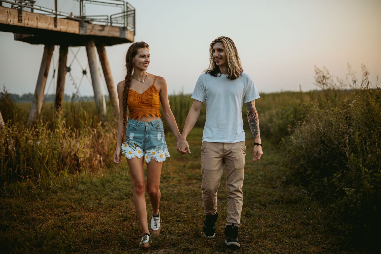 Couple walking hand in hand along path near metal structure during sunset.