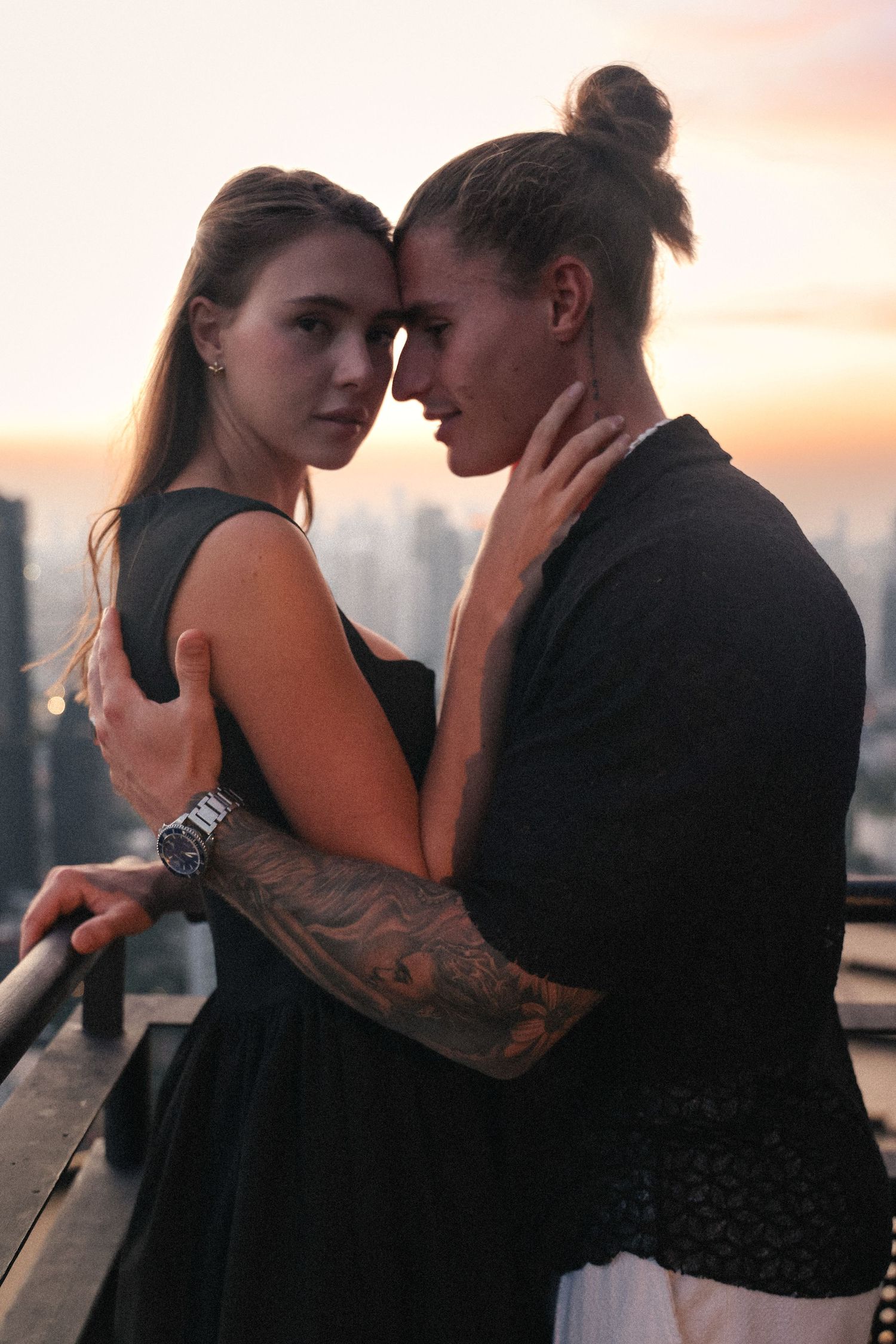 Intimate couple embracing on rooftop with city sunset in background.
